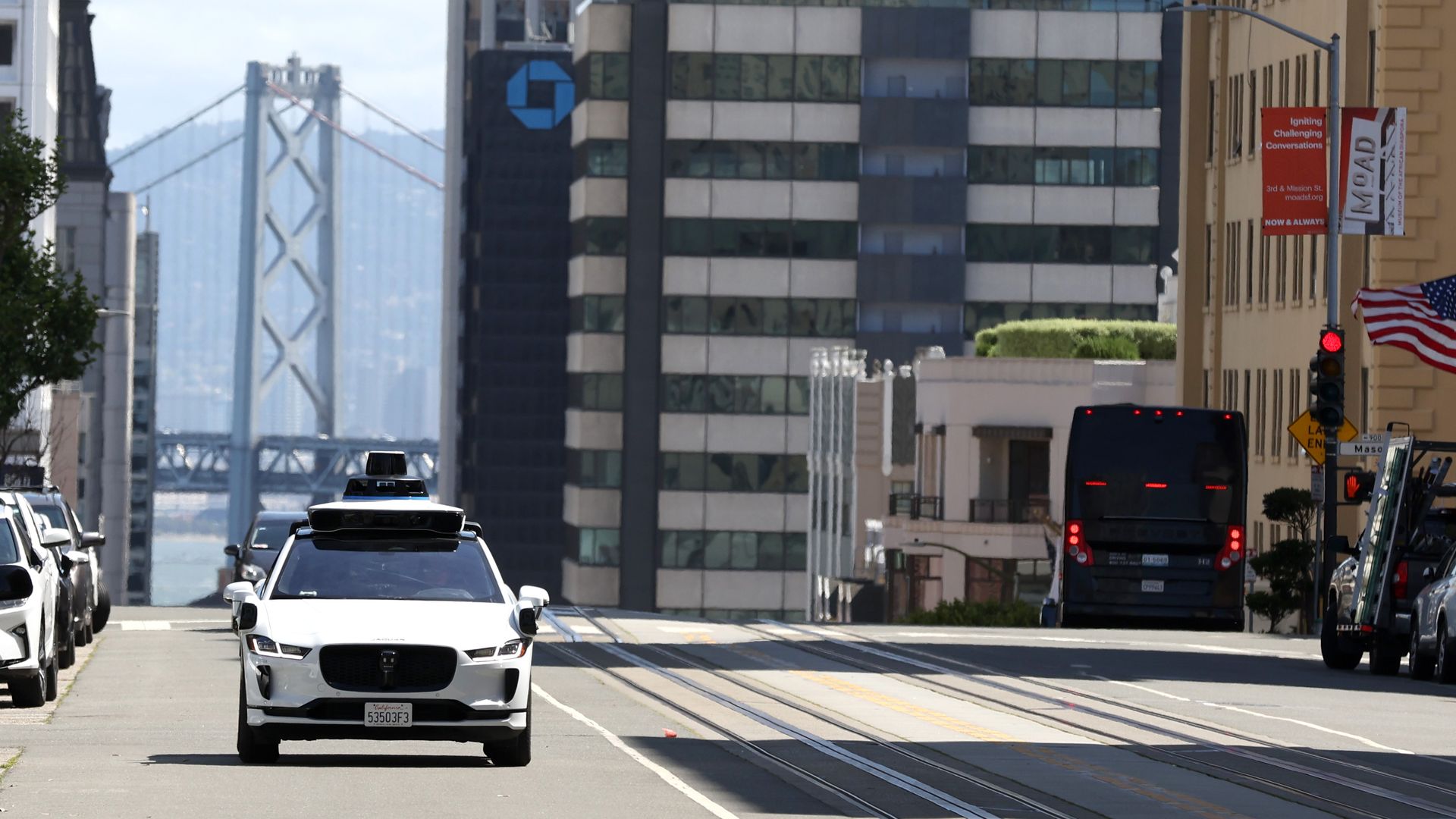 Man walking in front of a self-driving car with the bay bridge in the background.