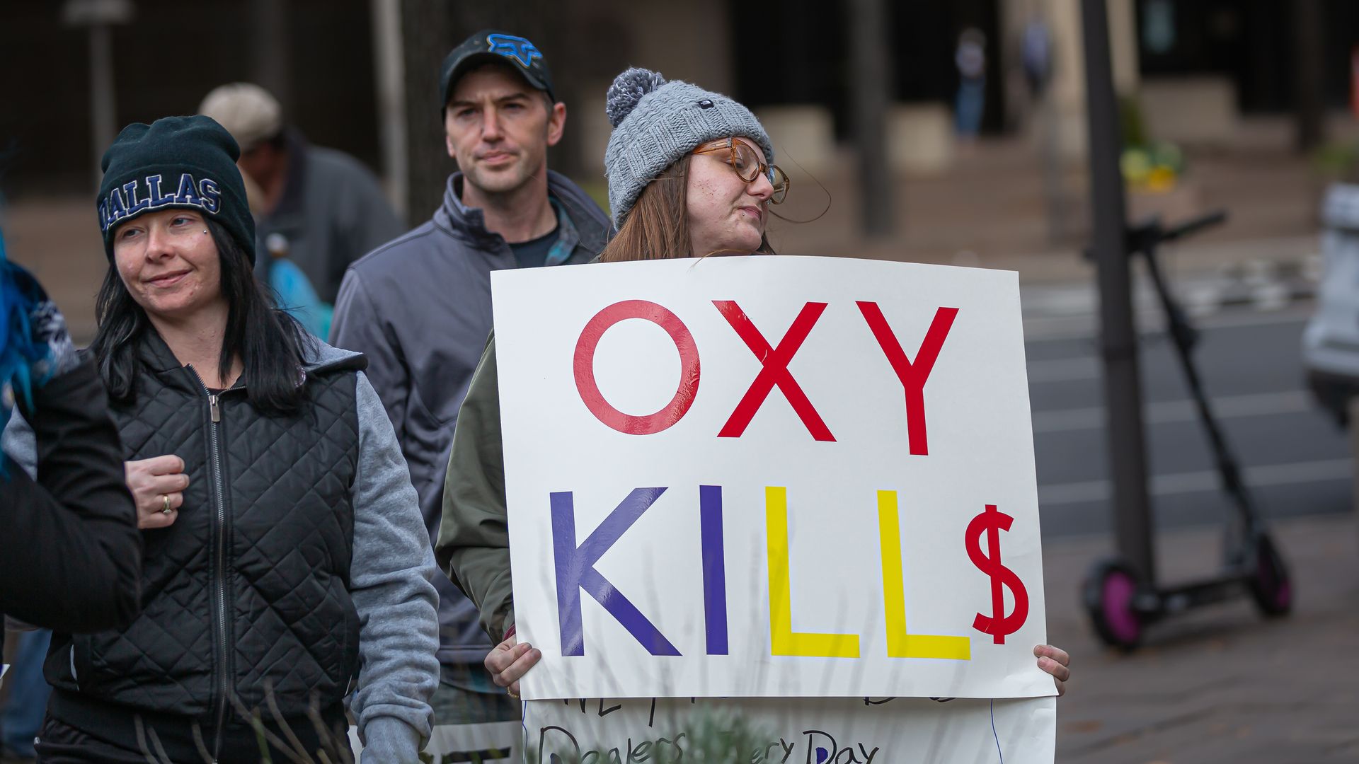 Picture of a person holding a sign that says "OXY KILLS" with a "$" instead of an "S" at the end