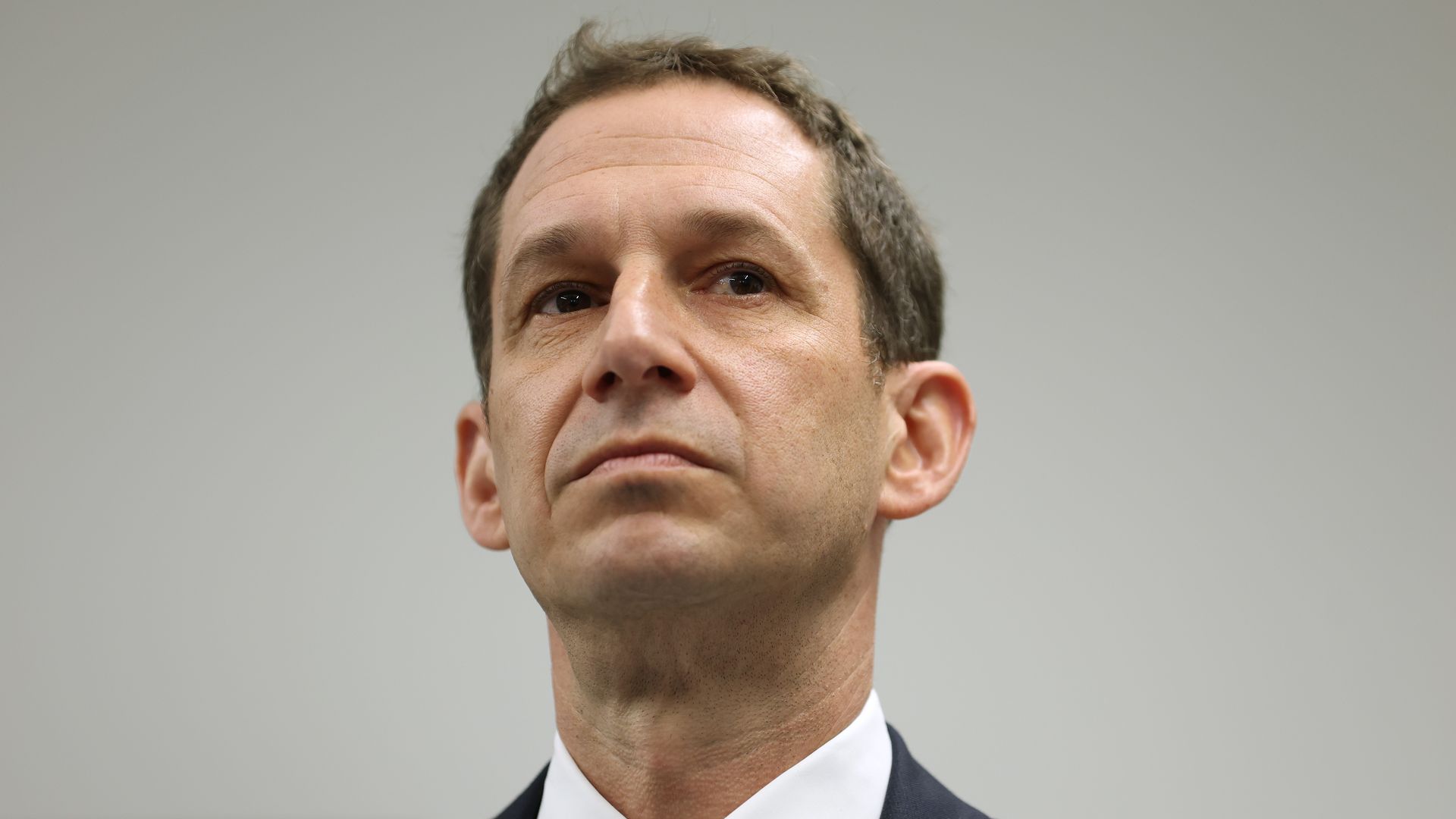 A middle-aged man in a dark suit and white shirt with a red patterned tie, looking slightly upward with a neutral expression, against a plain gray background.