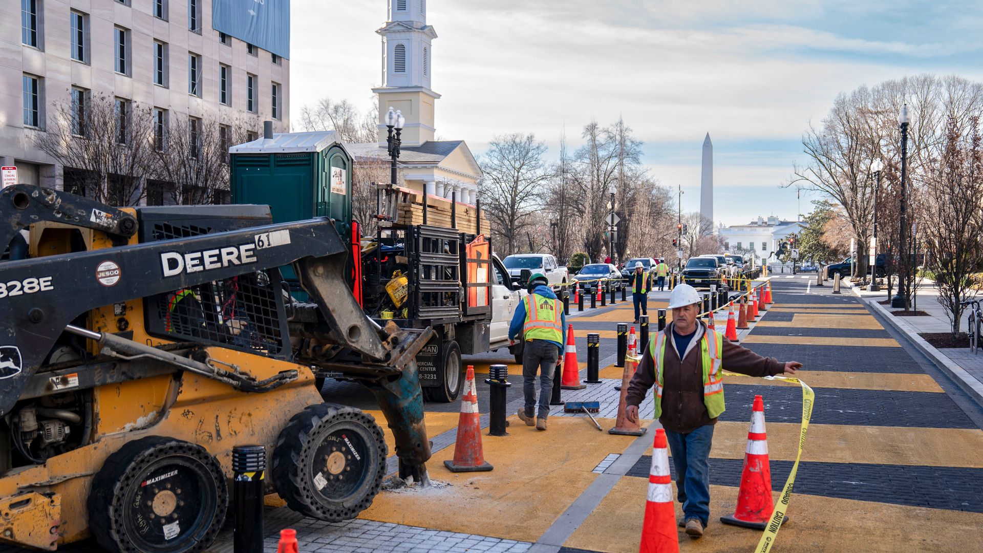 Construction workers in safety vests and helmets work on a street with orange cones and machinery, with the Washington Monument and government buildings visible in the background under a cloudy sky.