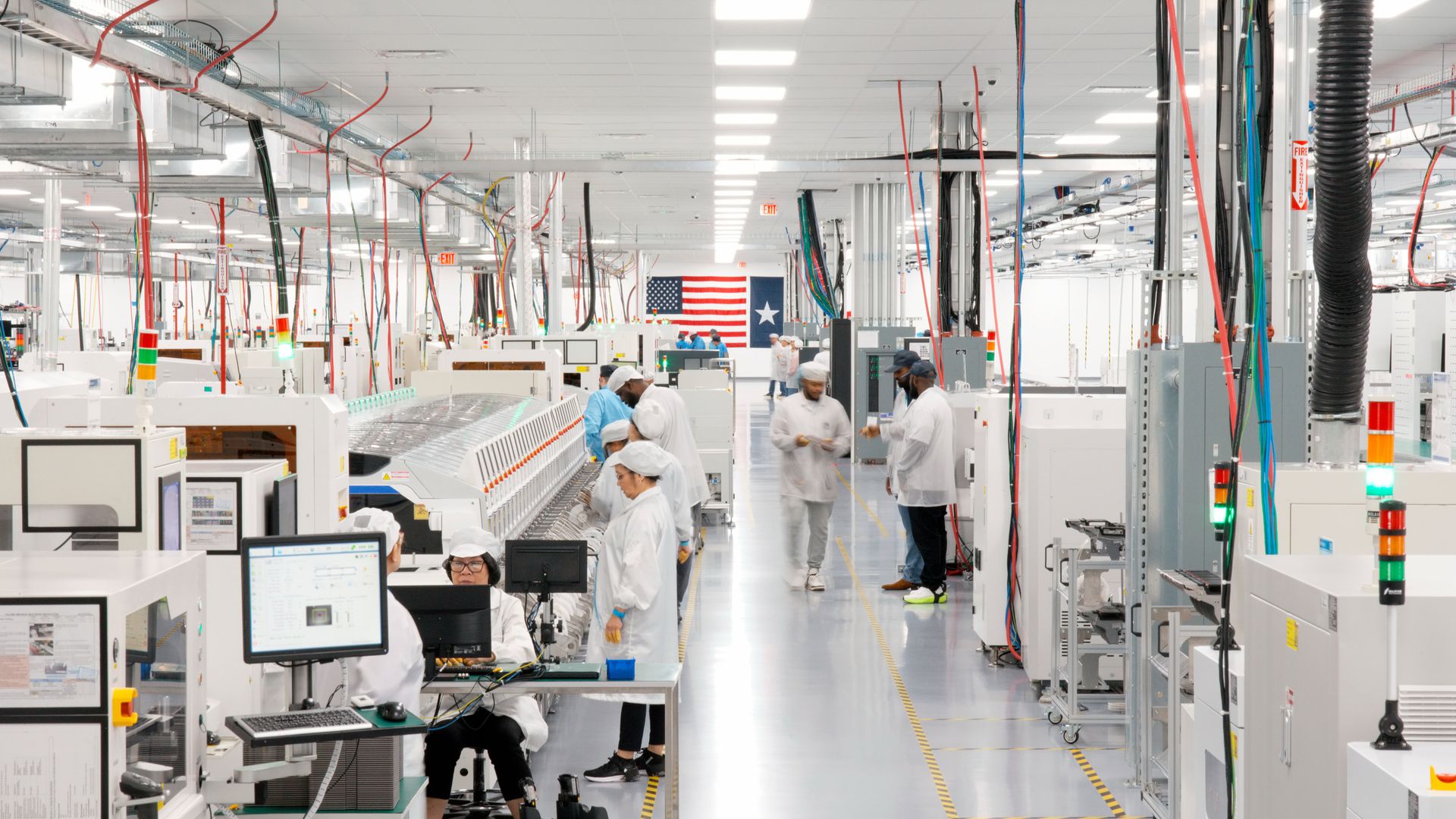 Bright, clean electronics manufacturing facility with workers in white lab coats and caps, operating machines and computers, American and Texas flags in background.
