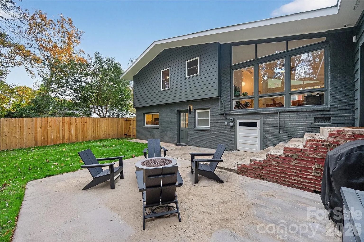 Backyard with gray two-story house, large windows, wooden fence, green lawn, concrete patio with four black Adirondack chairs around a round fire pit, and brick stairs.