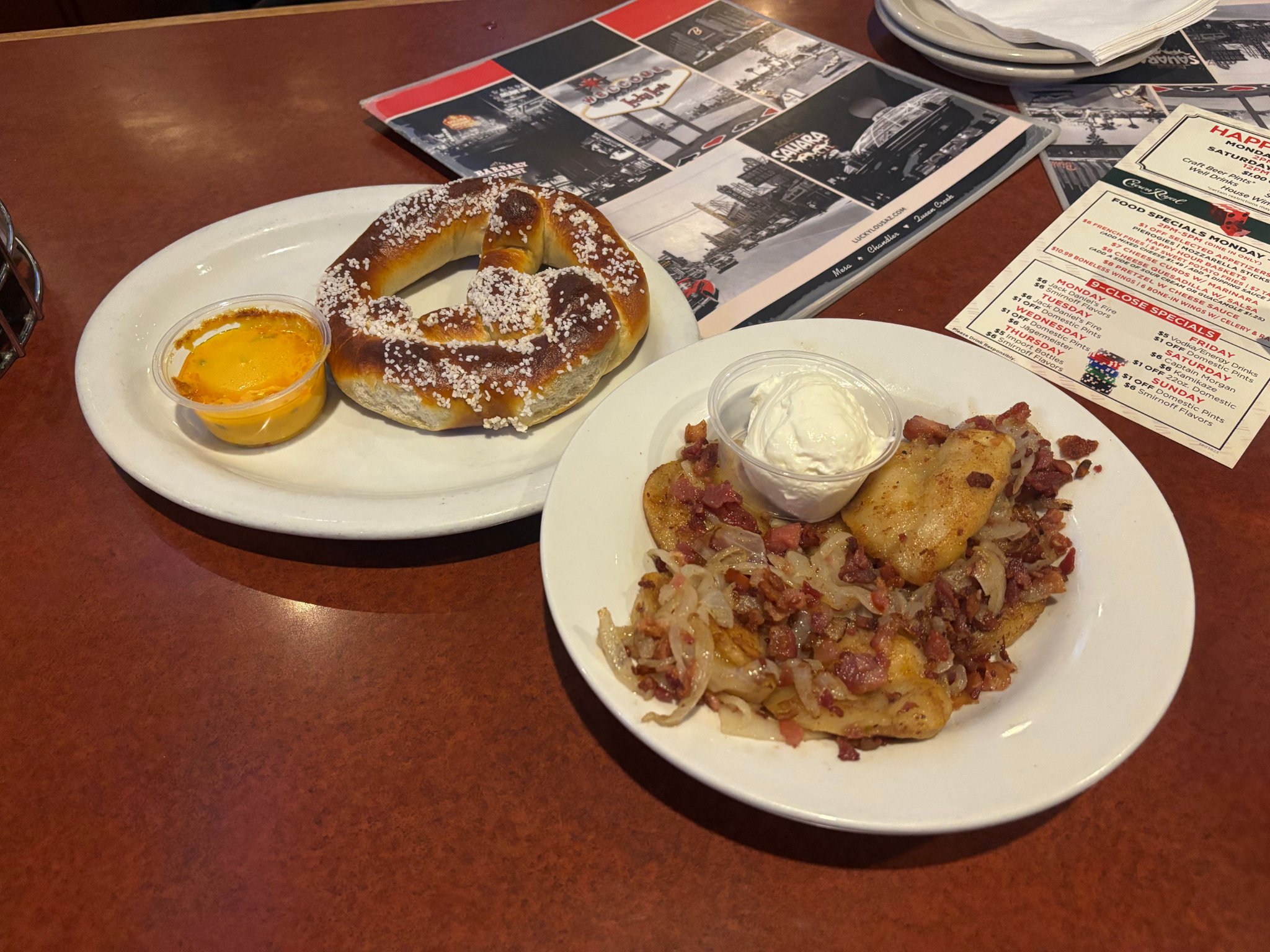 Close up on a diner table with two plates: One with a pretzel and cheese, the other with pierogis covered in onions and bacon with a side of sour cream.