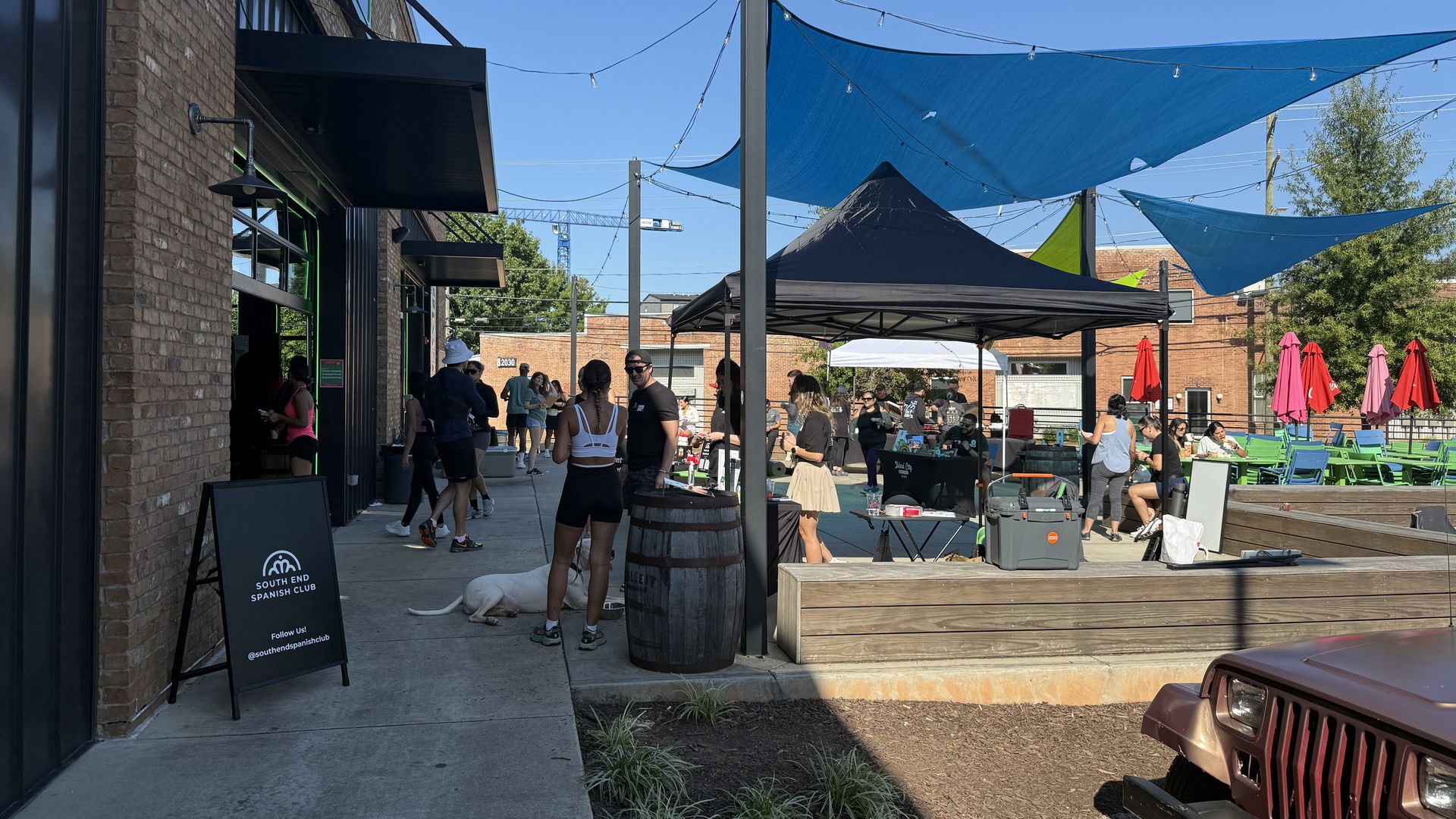 Outdoor market under blue shade sails with a brick building on the left, string lights, and groups of people near tents. A white dog lies on the ground beside a sign reading "South End Spanish Club".