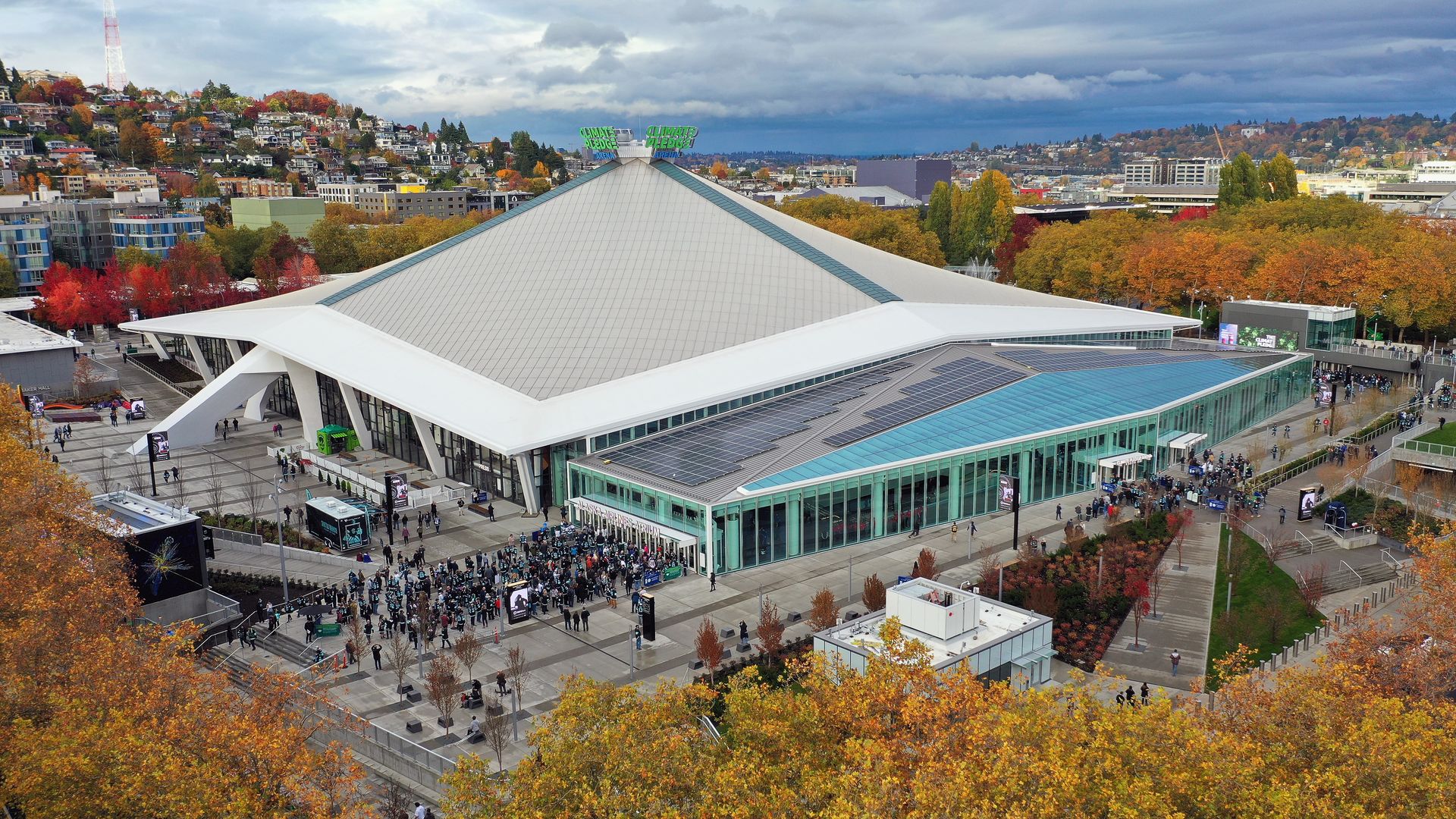 An aerial view of Climate Pledge Arena surrounded by trees and a view of part of Seattle, as people line up to enter the arena.