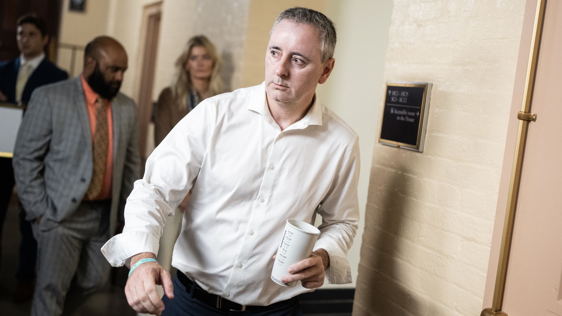 Rep. Brian Fitzpatrick, wearing a white shirt and blue pants and holding a white coffee cup, walks through a white brick basement hallway.