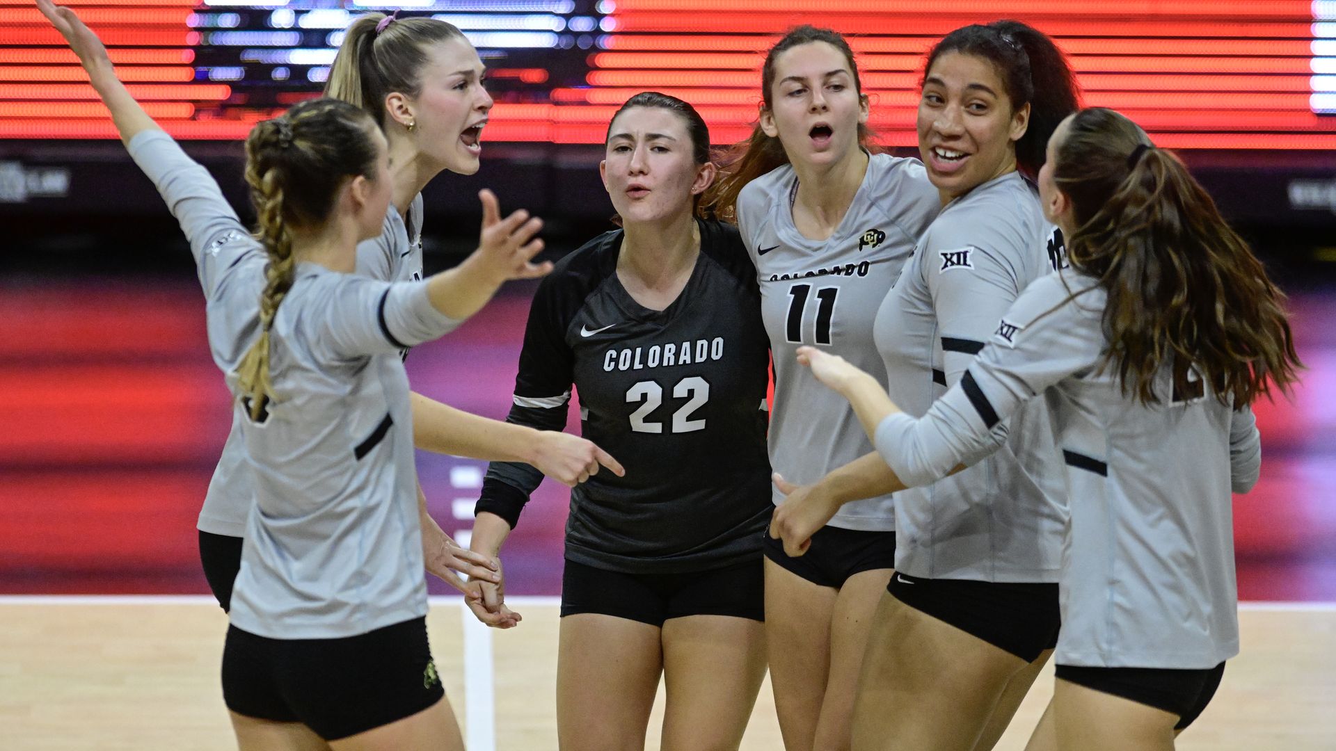 Six Colorado volleyball players on court in gray and black uniforms celebrating or discussing, with expressive faces and hand gestures, against a red digital backdrop.