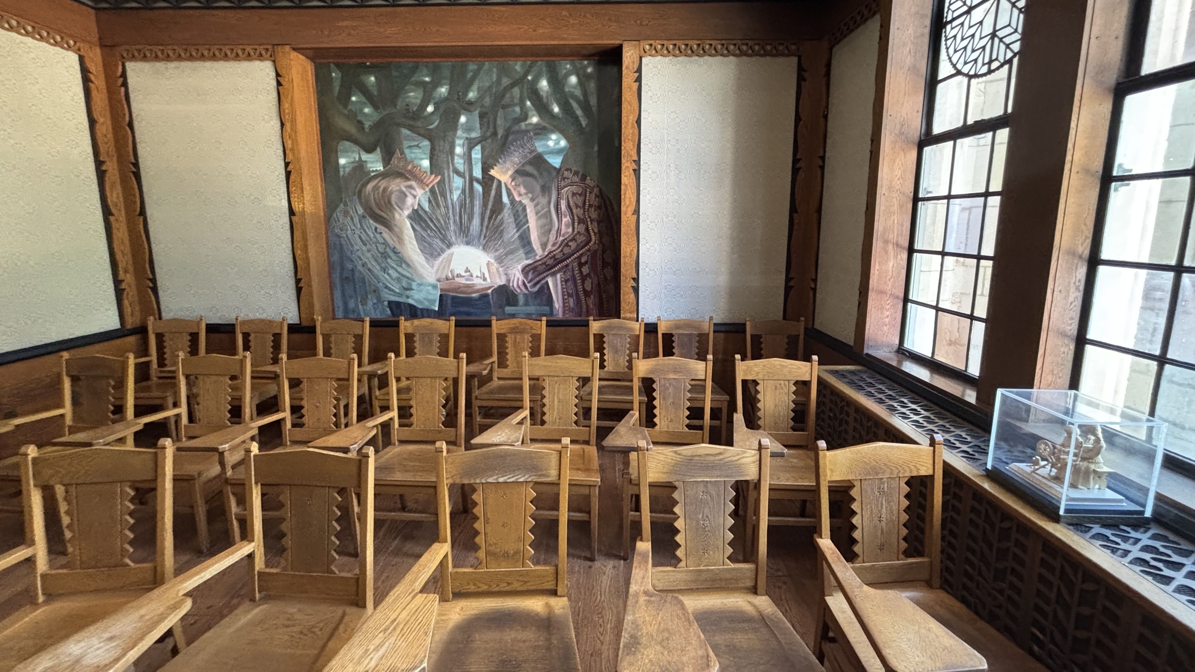 Wood-paneled room with rows of carved wooden chairs; mural at back shows two crowned figures around a glowing orb among dark trees; windows along the right wall.