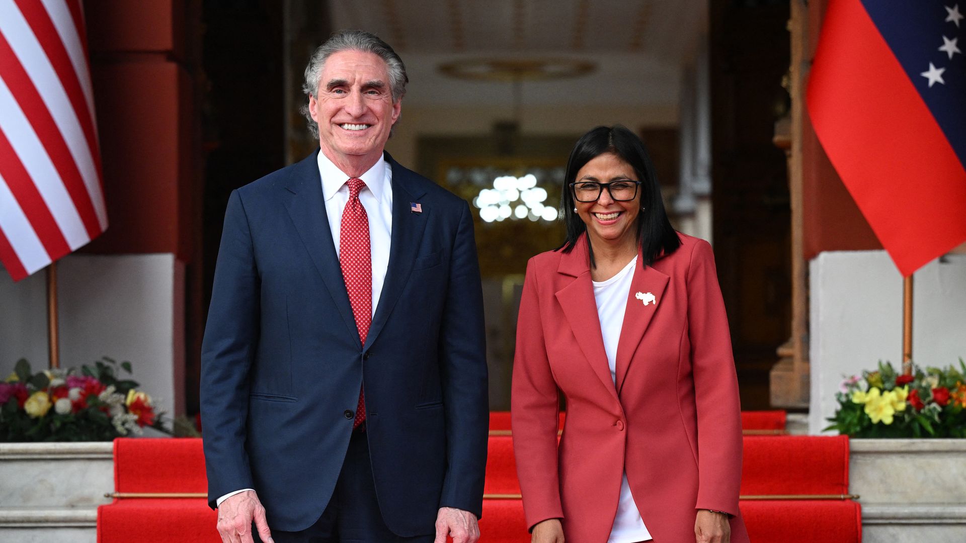 Interior Secretary Doug Burgum and Venezuela's interim president, Delcy Rodriguez stand smiling on red carpeted steps between U.S. and Venezuelan flags; Burgum wears navy suit red tie, Rodriguez wears red suit with white shirt and glasses.