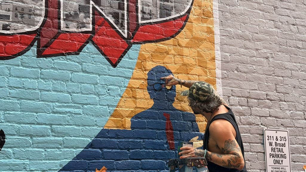 Graffiti mural on a brick wall in red, blue, and yellow. A man wearing a camouflage cap and sleeveless shirt paints a blue silhouette, holding a palette; a parking sign sits to the right.