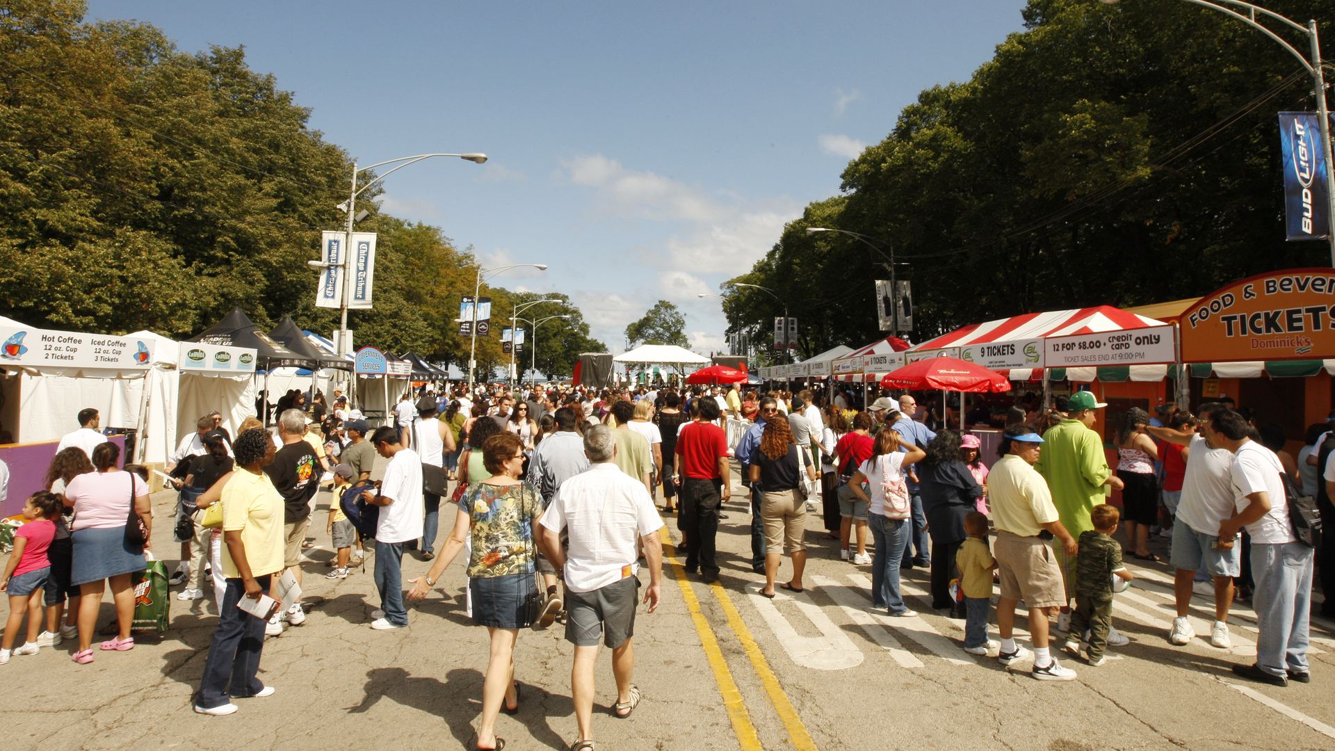 Busy outdoor festival on a sunny day with a crowded street lined by white vendor tents, blue banners, and tall green trees, under a clear blue sky.