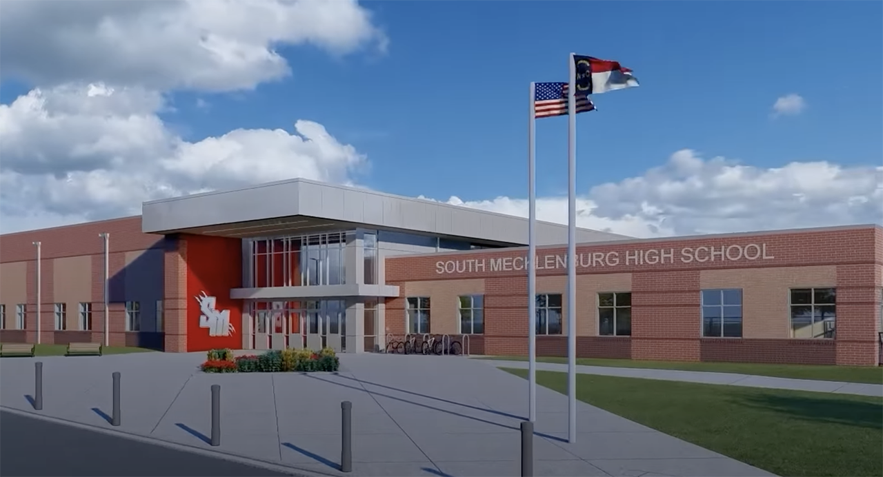Front of South Mecklenburg High School, a brick building with a glass entrance, red wall logo, and two flagpoles flying the U.S. and North Carolina flags under a blue sky with clouds.