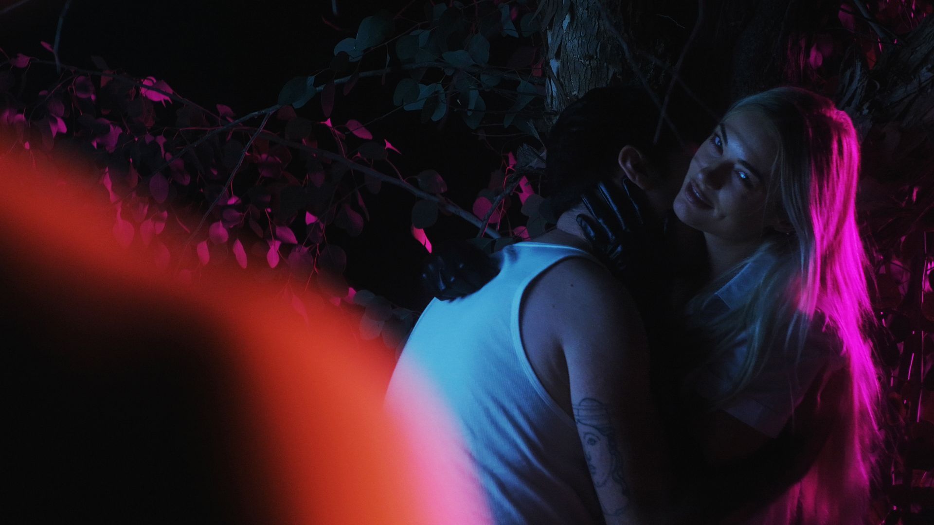 Two people in a dark outdoor scene lit by pink and blue neon lights; a smiling woman with long hair in a white sleeveless top faces the camera, while another person stands nearby among pink foliage.