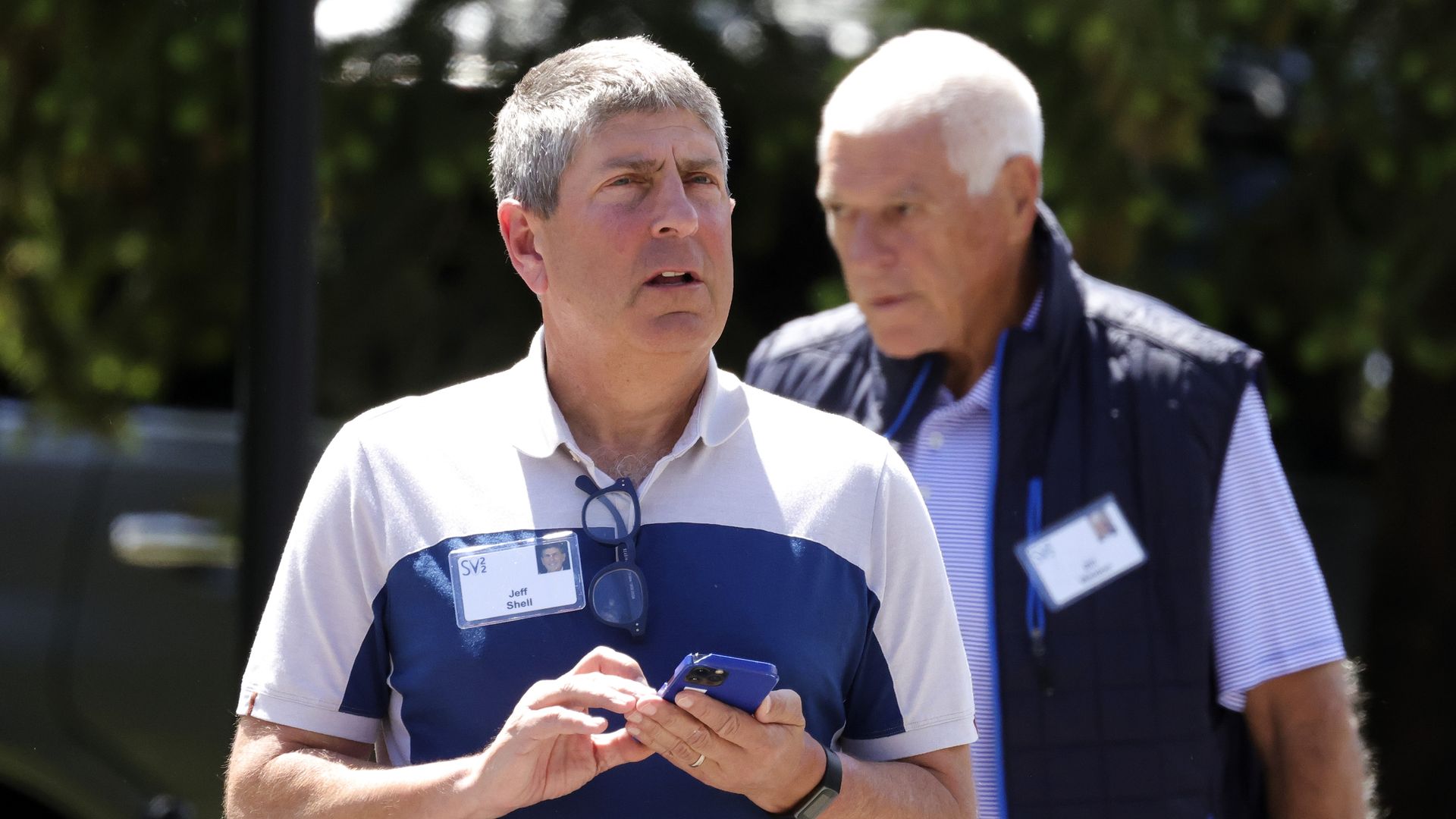Jeff Shell stands outdoors in sunlight, holding a smartphone and looking off to the side with a concerned expression. He wears a white and blue polo shirt with a name badge clipped to it, and sunglasses hanging from the collar.