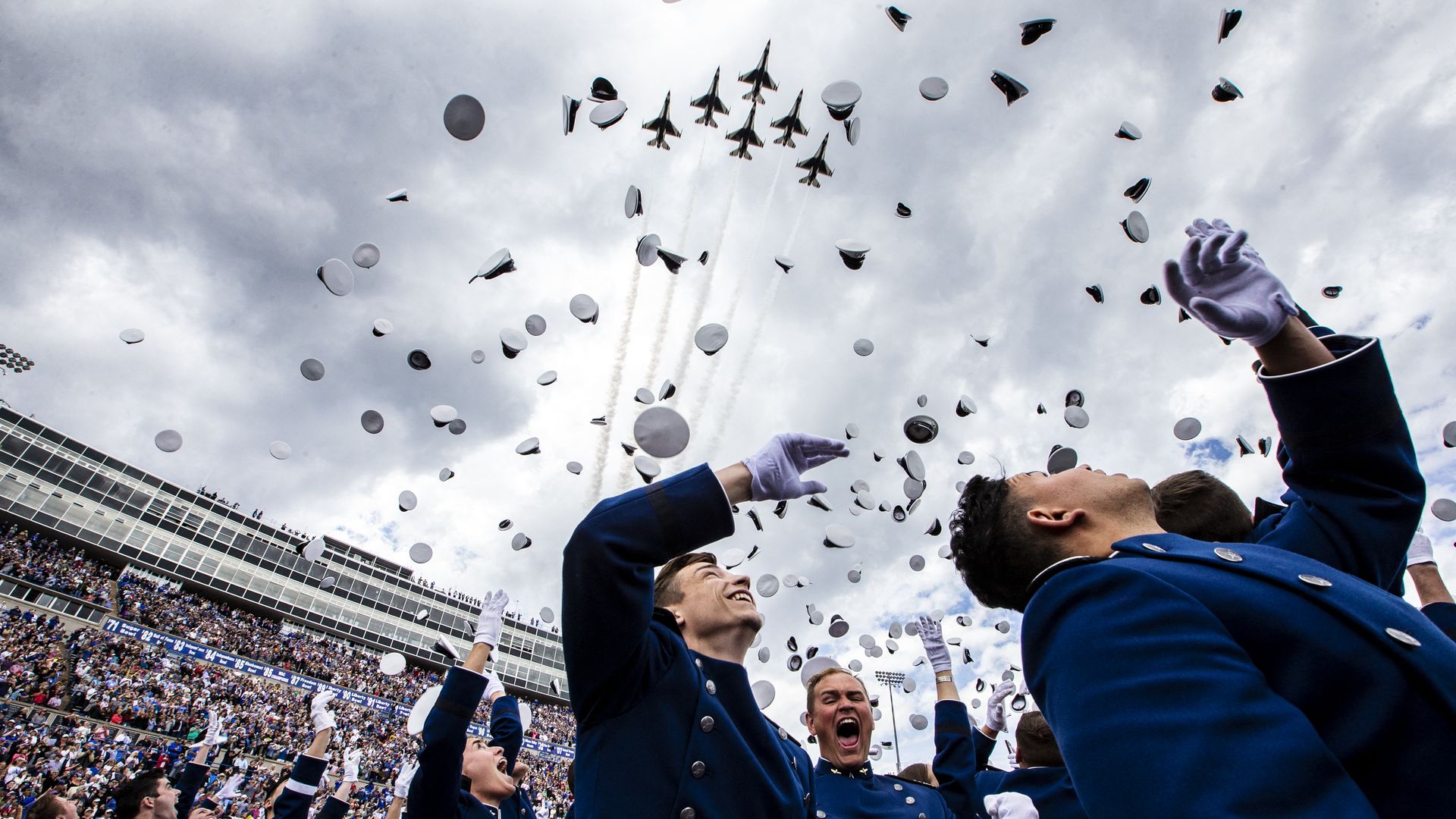 People in blue military uniforms smile and cheer as they throw white caps into the sky. The sky is cloudy, and six jets in tight formation can be seen streaking overhead. 