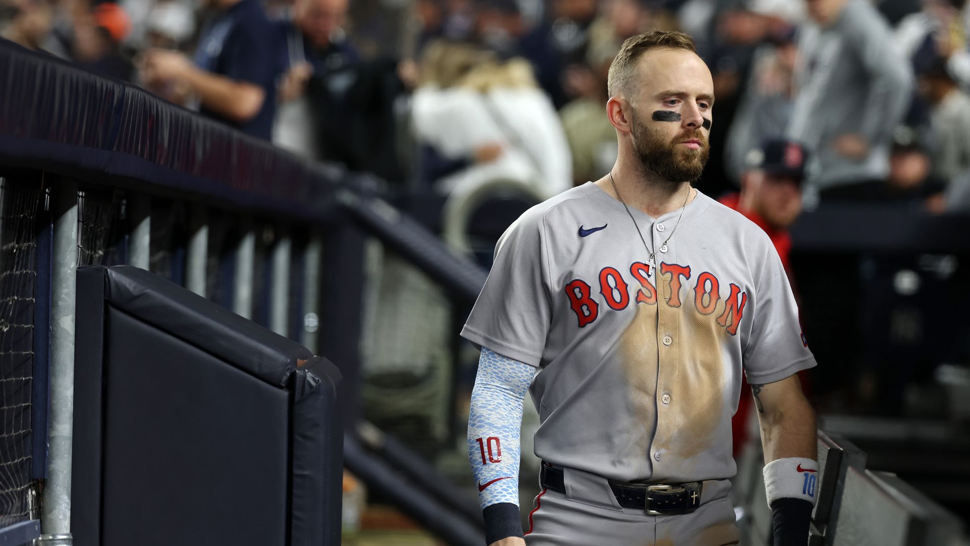 Baseball player in a dirty gray Boston jersey with black eye paint, walking near dugout during game, wearing blue arm sleeve and white wristband, focused expression, crowd blurred in background.