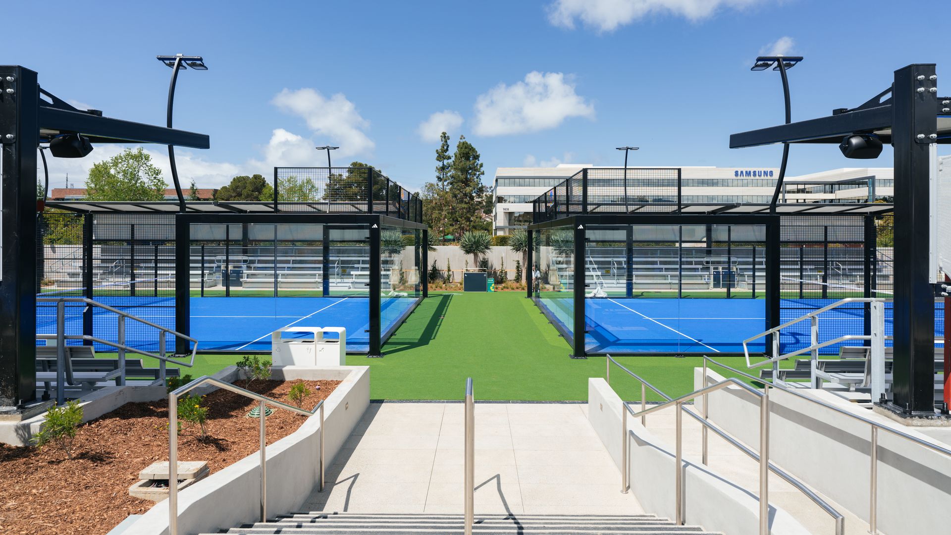 Modern outdoor sports courts with blue playing surfaces, black metal fencing, and surrounding green turf under a blue sky with scattered clouds, with a Samsung building visible in the background.