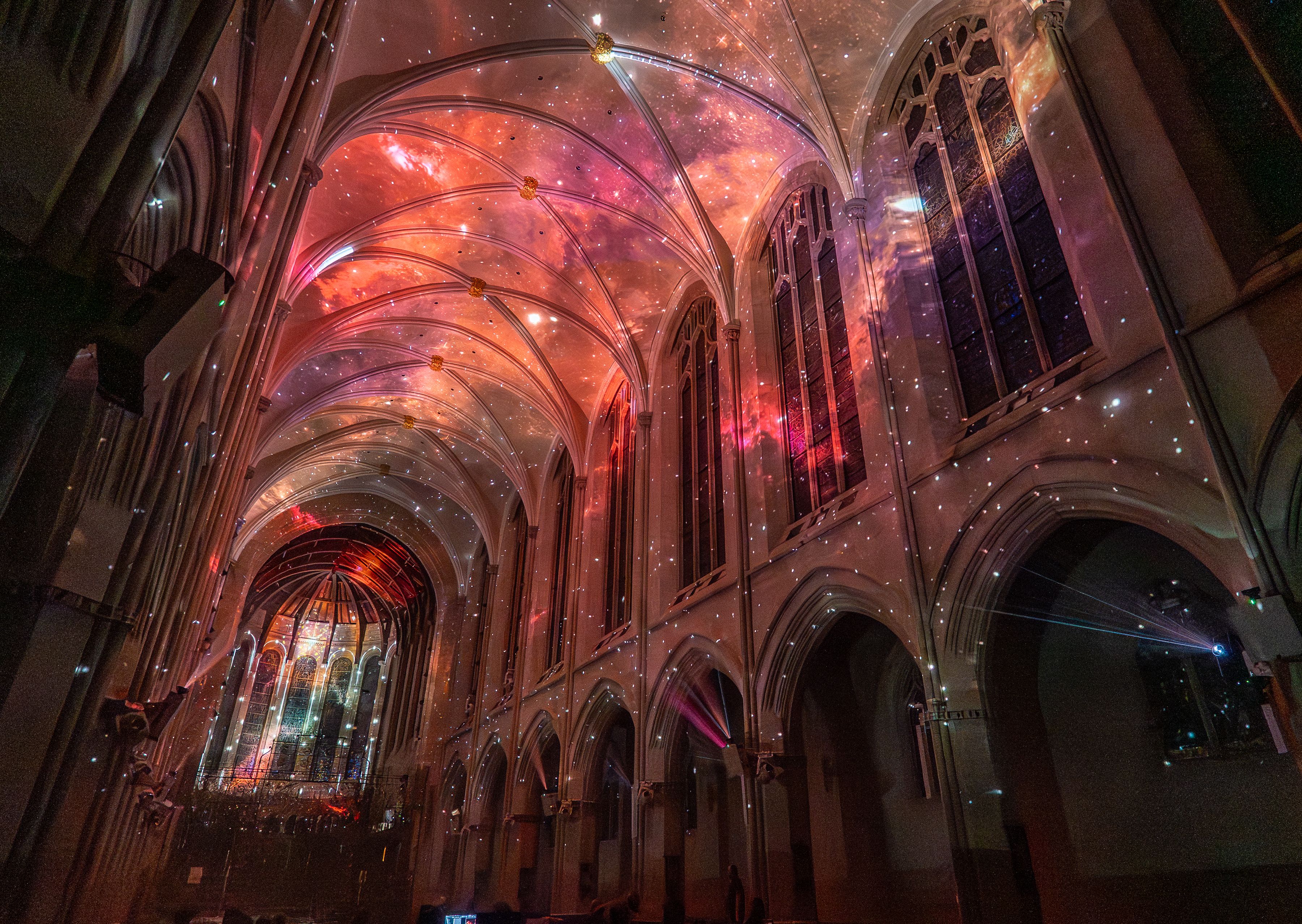 Inside a Gothic cathedral with white ribbed arches, warm pink-orange galaxy lights on the ceiling, a starry sky projection, and tall stained-glass windows along the sides.