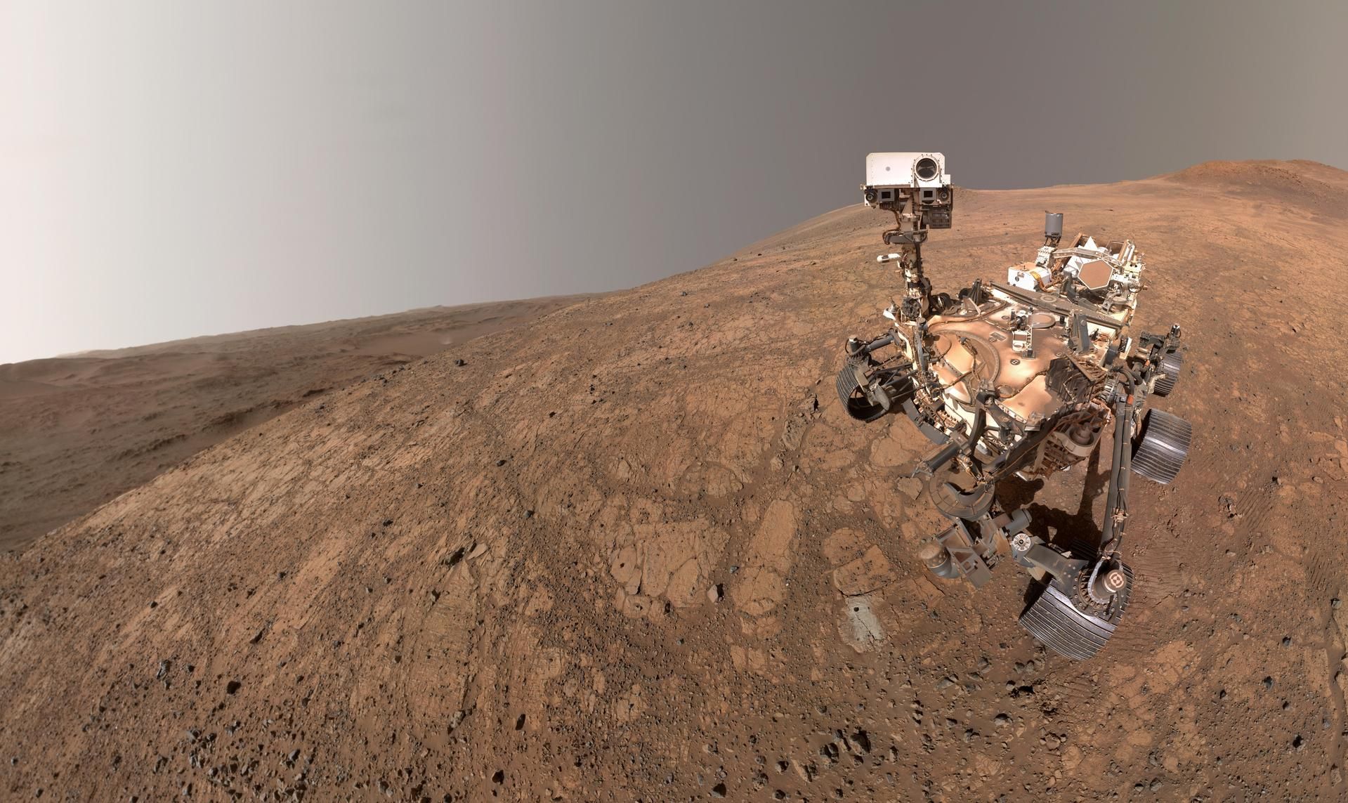 Mars rover on orange-red, rocky terrain with a camera mast and robotic arm; large wheels and instruments visible against a pale gradient sky.