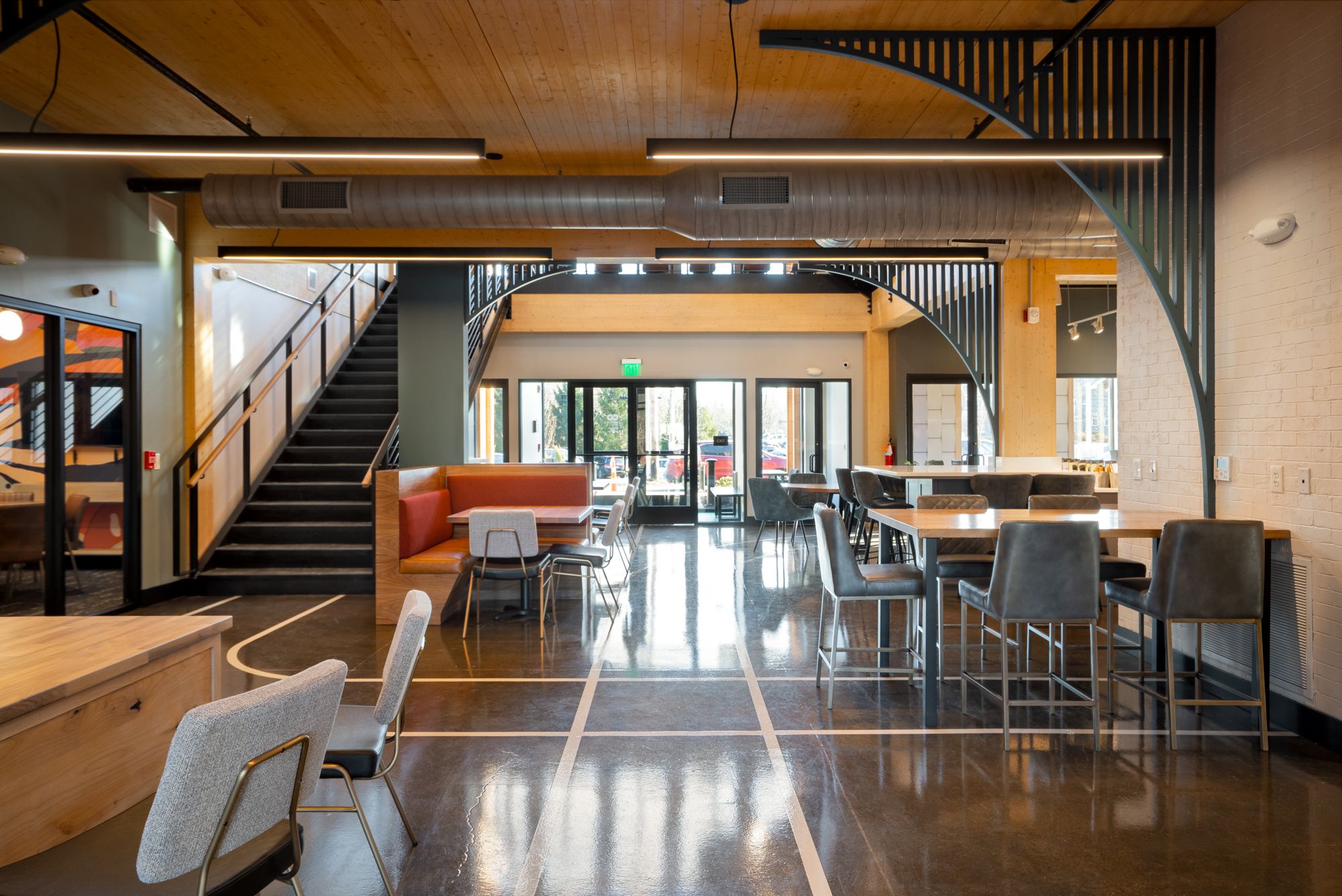 Modern indoor lounge area with polished concrete floors, wooden ceiling, black staircase, gray and orange seating, high tables with gray chairs, and large windows letting in daylight.