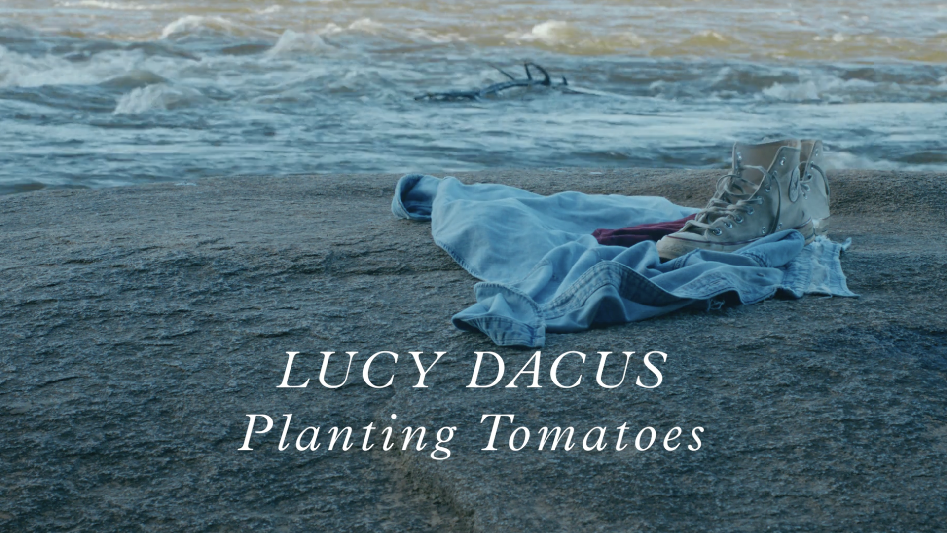 A light blue blanket and tan boots lie on a rocky beach with blue waves; bottom text reads "LUCY DACUS Planting Tomatoes" in white script.