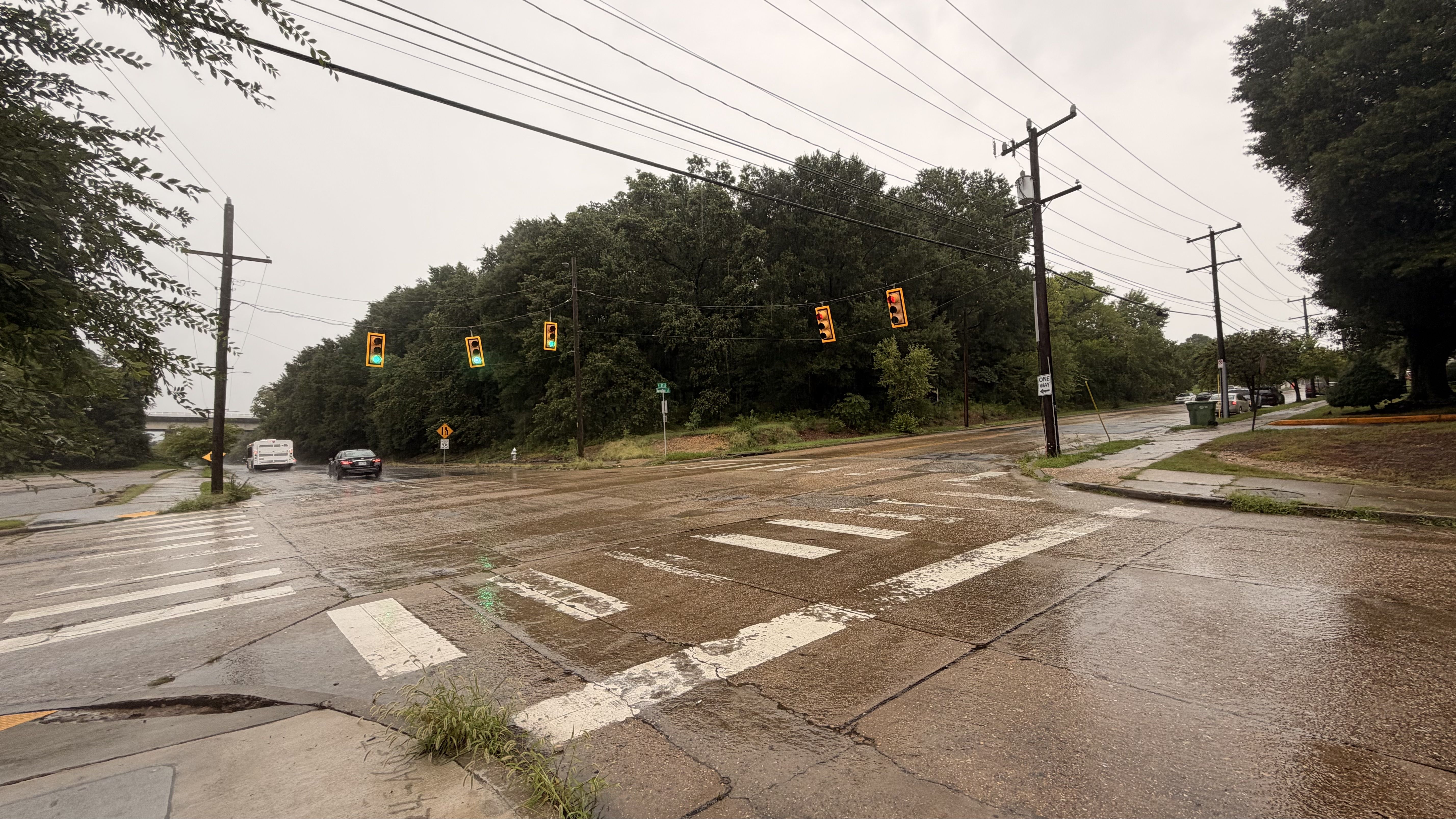 Rainy daytime at a wide street intersection: traffic lights strung across the wet road, crosswalk markings, utility poles and overhead wires, with trees to the right and a white van and black car.