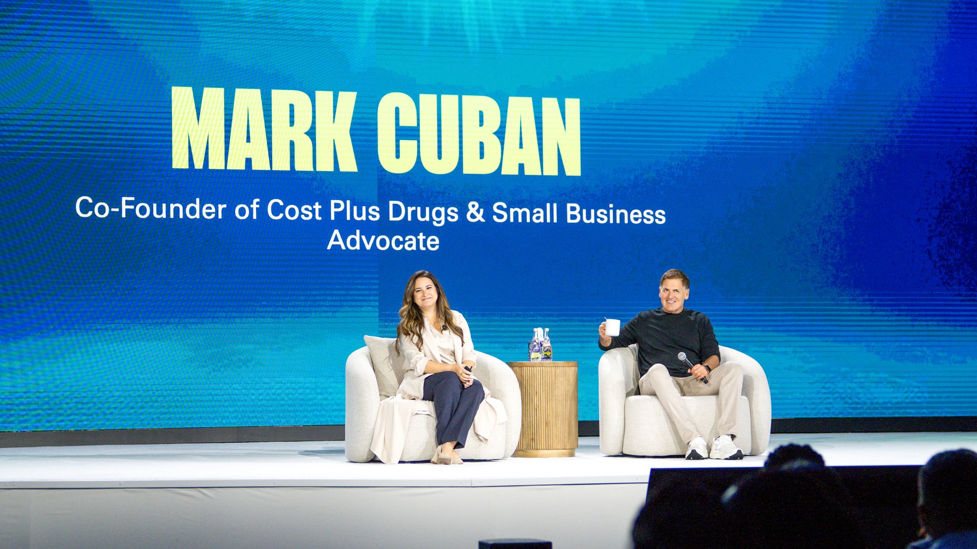 Mark Cuban and a woman seated on beige armchairs on a stage with a blue background displaying his name and titles: Co-Founder of Cost Plus Drugs & Small Business Advocate.