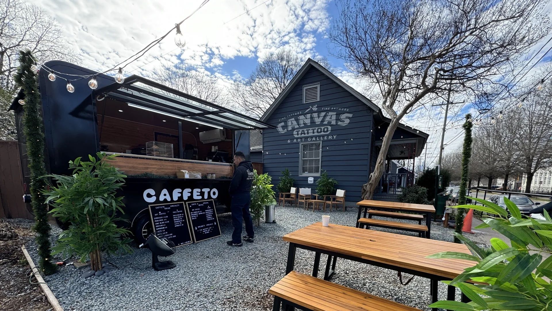 Outdoor café seating with wooden tables and benches on gravel near a black food truck labeled "CAFFETO" and a blue building with "CANVAS Tattoo & Art Gallery" sign, under a partly cloudy sky.