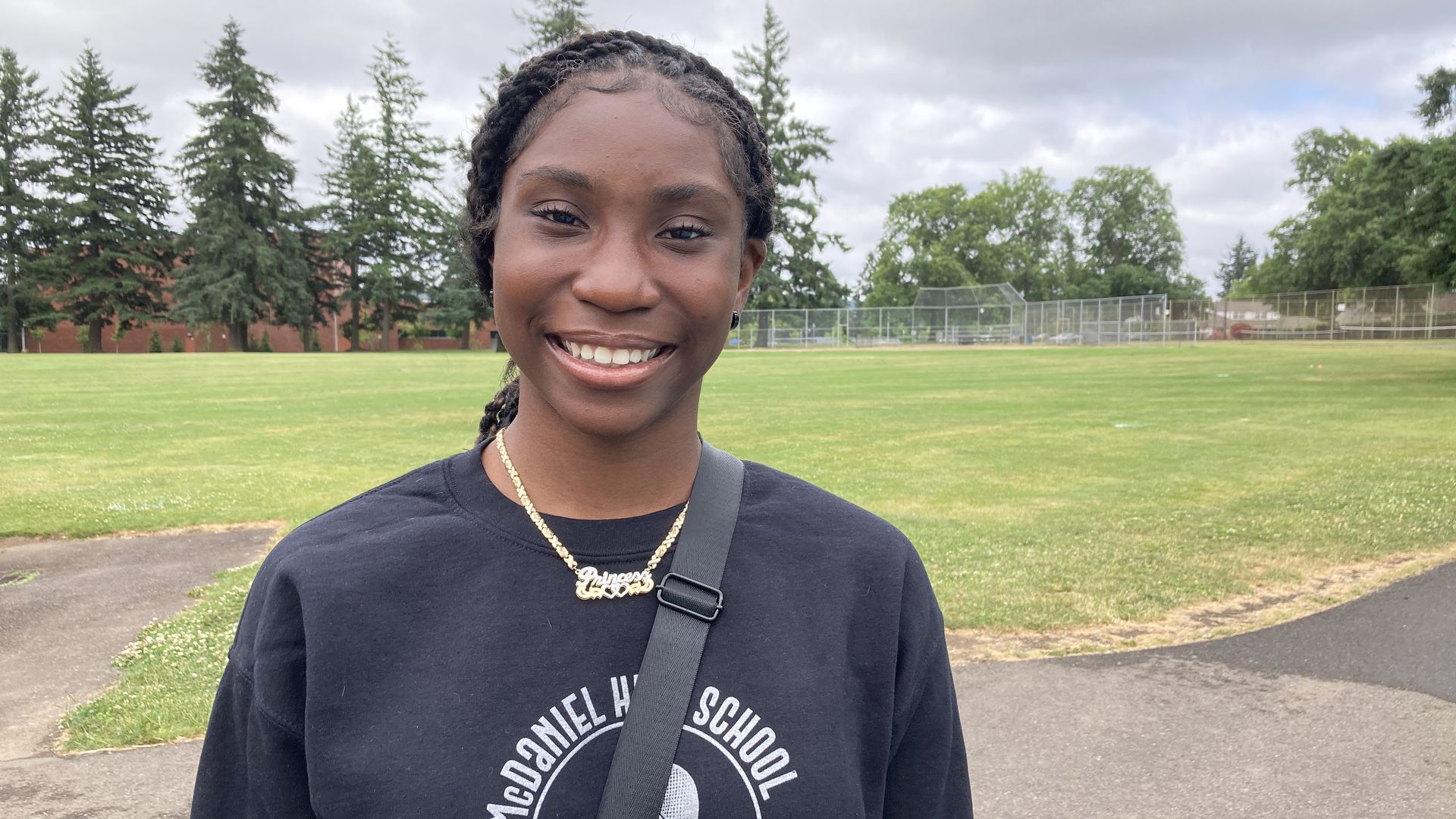 A smiling young woman with a gold necklace that says Princess and a blue sweatshirt with a high school emblem.