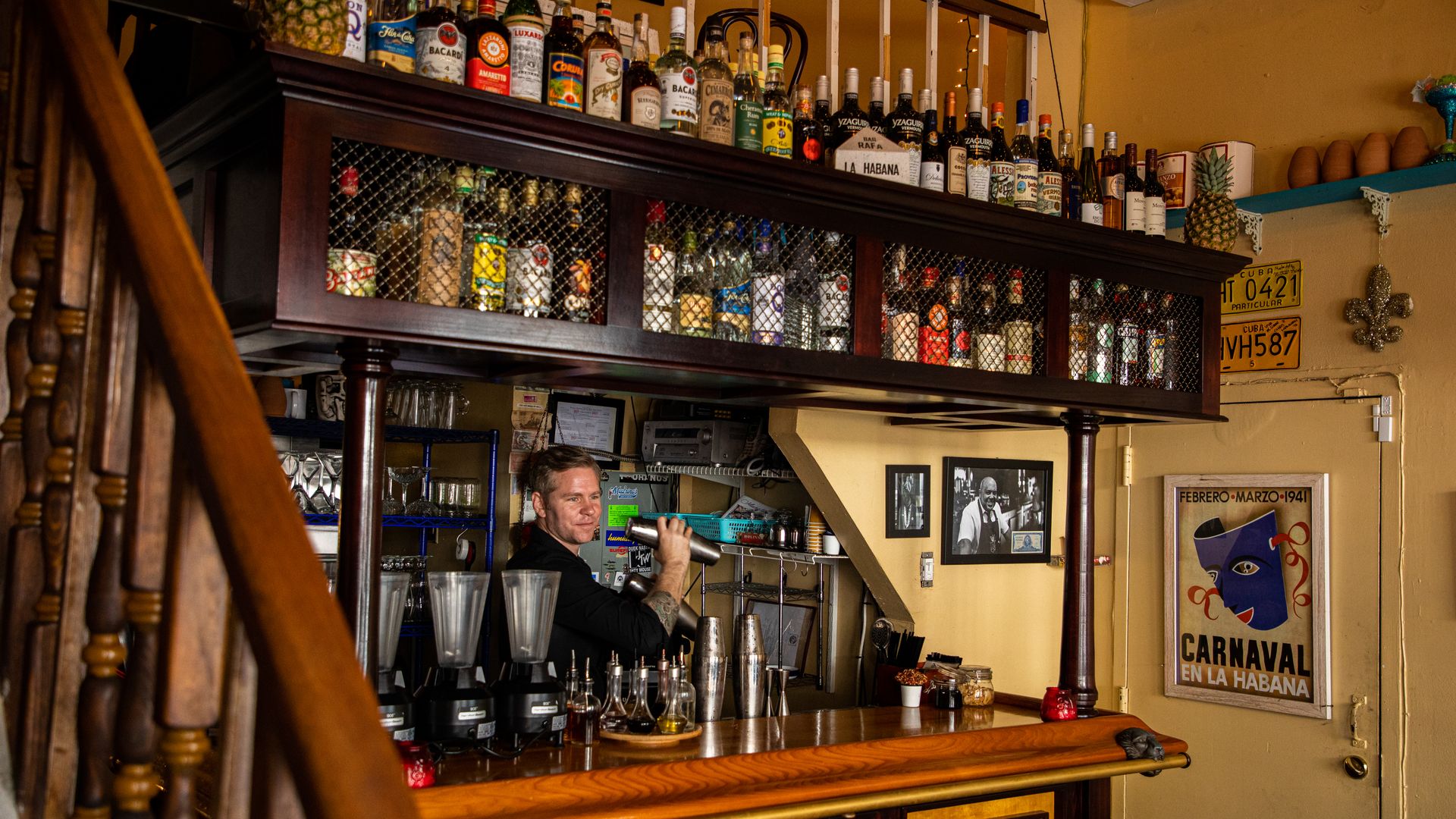 A bartender shakes a cocktail inside the Manolito bar. Above the bar, two rows of liquor bottles sit on shelves with their labels turned neatly outward.