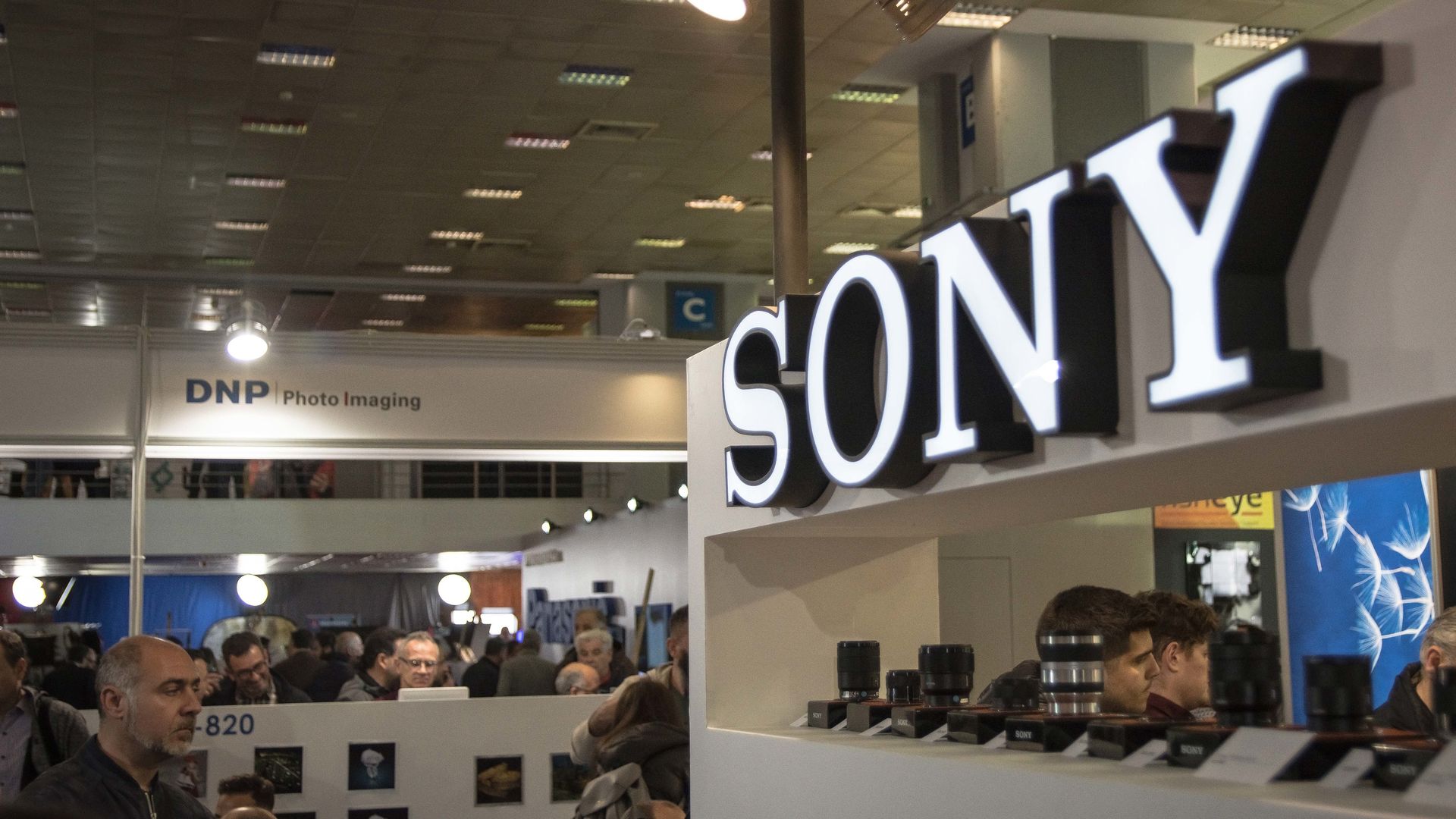 The Sony logo is seen in white on a wall as people walk around and look at phones and other tech.