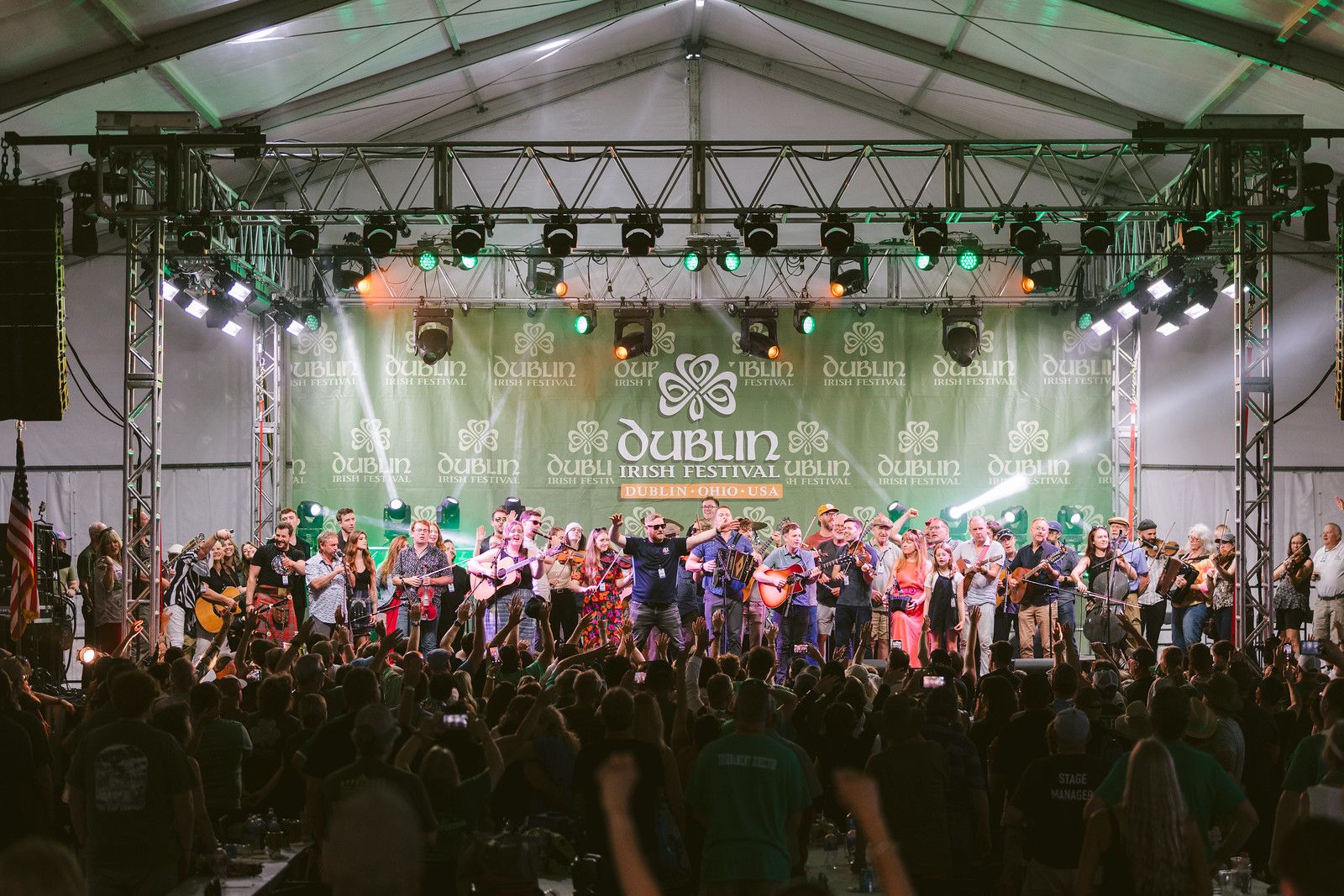 Stage under a white tent with green lights; a large group of musicians and singers play guitars, fiddles, and accordions as a crowd watches. Backdrop reads "Dublin Irish Festival".