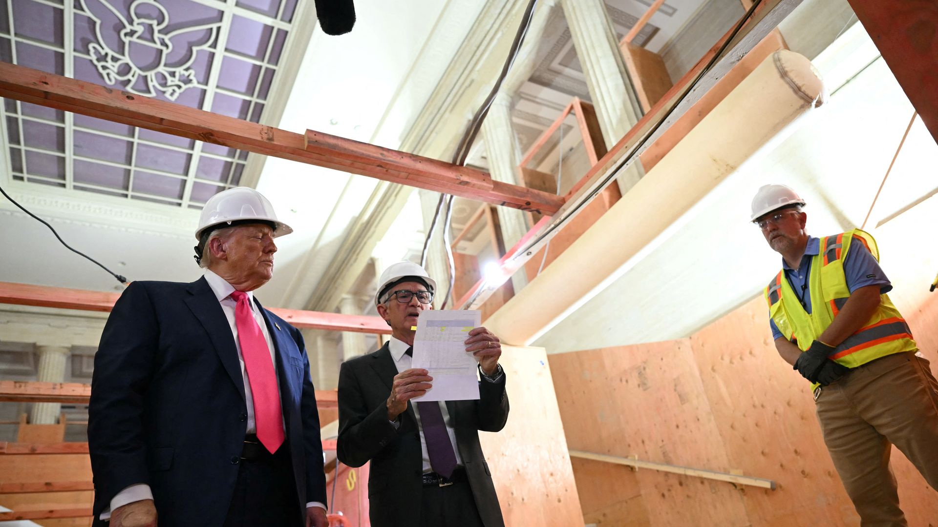 Three men wearing hard hats at a construction site inside a building with high ceilings and columns; one man in a suit with a pink tie, another in a suit reading papers, and a worker in a safety vest.