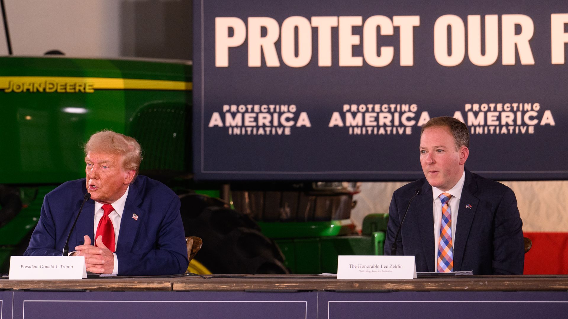  President Trump, wearing a navy jacket with a US flag pin at the top of his left lapel, white shirt and red tie, and EPA chief Lee Zeldin, wearing a black jacket, white shirt and pink, blue and white tartan-style check tie, sit at a desk with their names on it in front of a blue and white sign.
