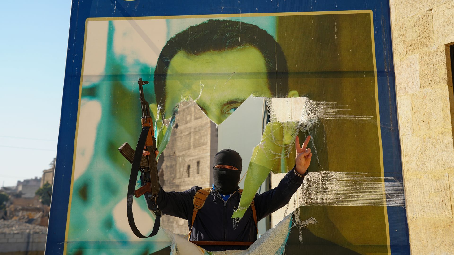 A person holding a gun busts through a large poster depicting Bashar al-Assad.