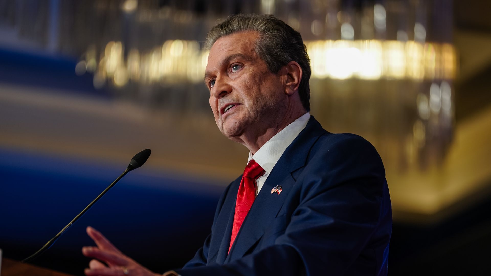 A man dressed in a dark-blue suit, red tie and white collared shirt speaks at a podium with a microphone. 