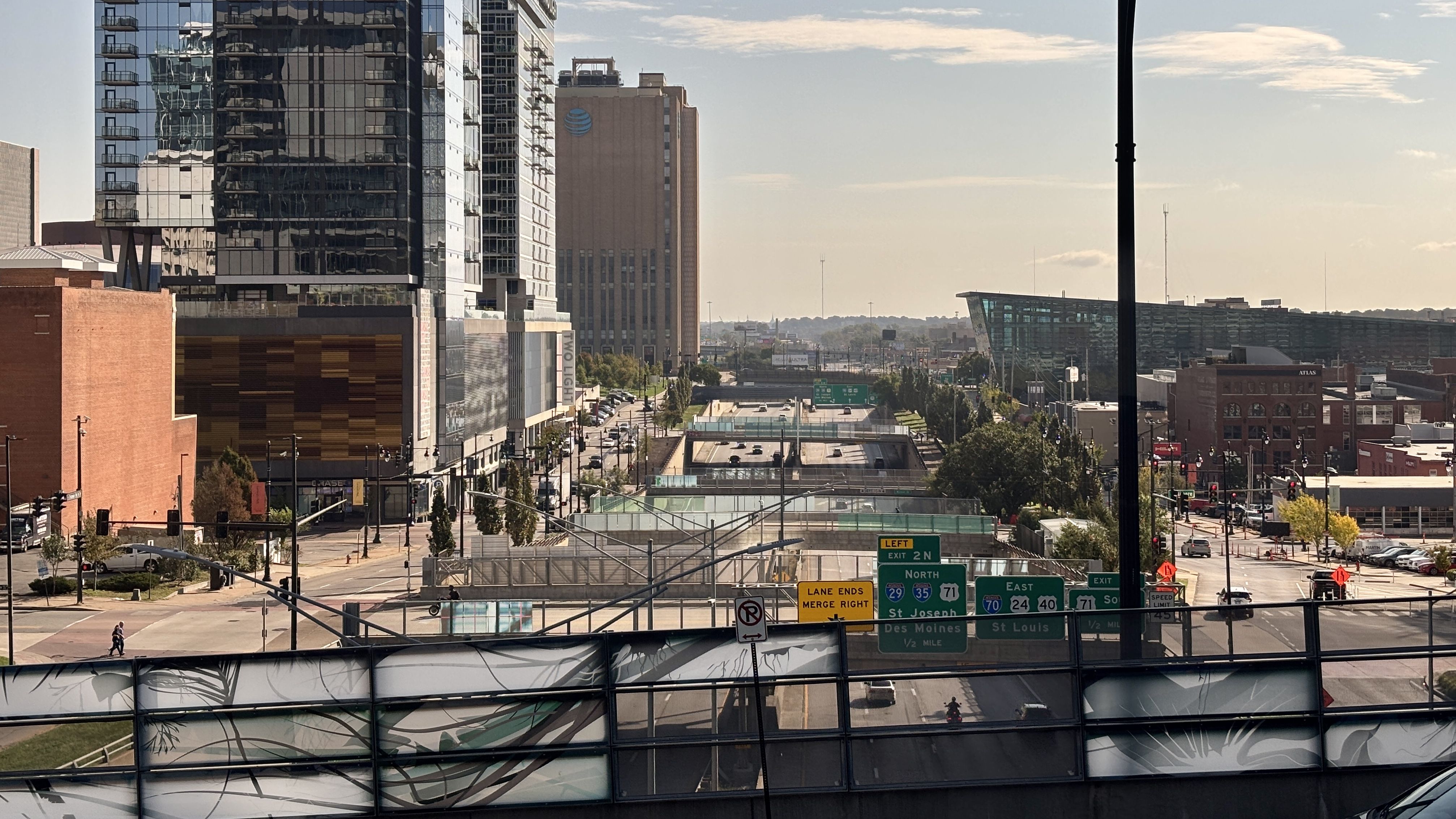 Urban cityscape seen from a bridge: glass high-rise on the left, tan AT&T-branded tower, a slanted glass building far right; busy multi-lane roads, highway signs, and a tall black streetlight against a pale sky.