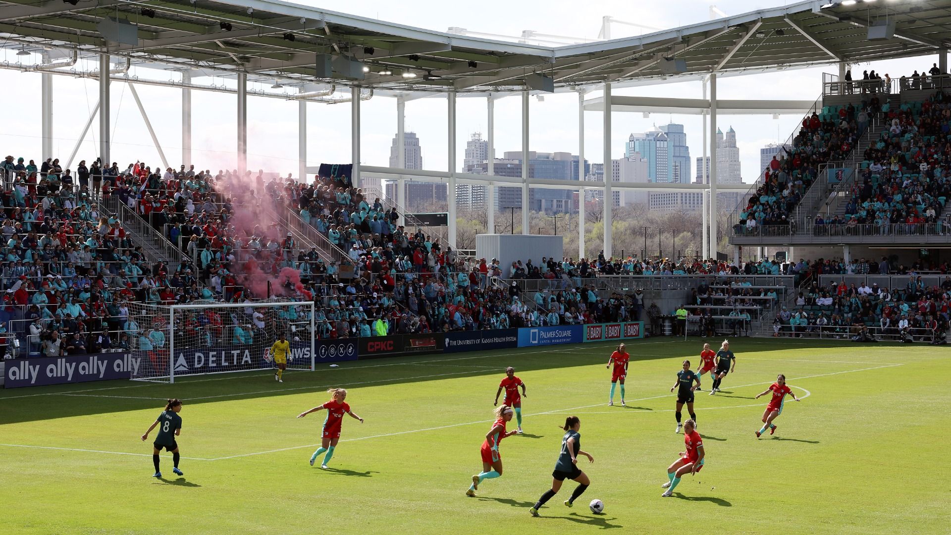Soccer match with players in red and dark teal uniforms on green grass field, large crowd in stadium seats, pink smoke flare in stands, city skyline in background.