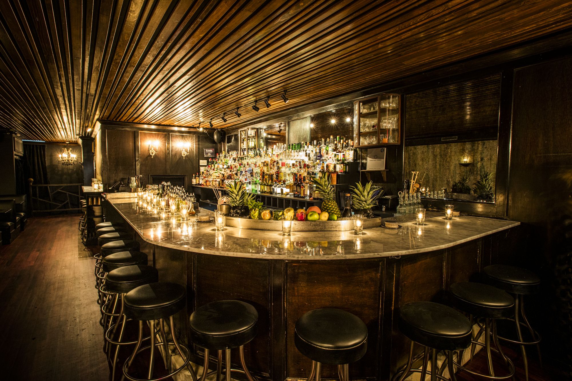 Warm, dimly lit bar with a marble countertop, surrounded by black stools. Shelves behind the bar hold various liquor bottles, fruits like pineapples and apples, and lit candles provide cozy ambiance.