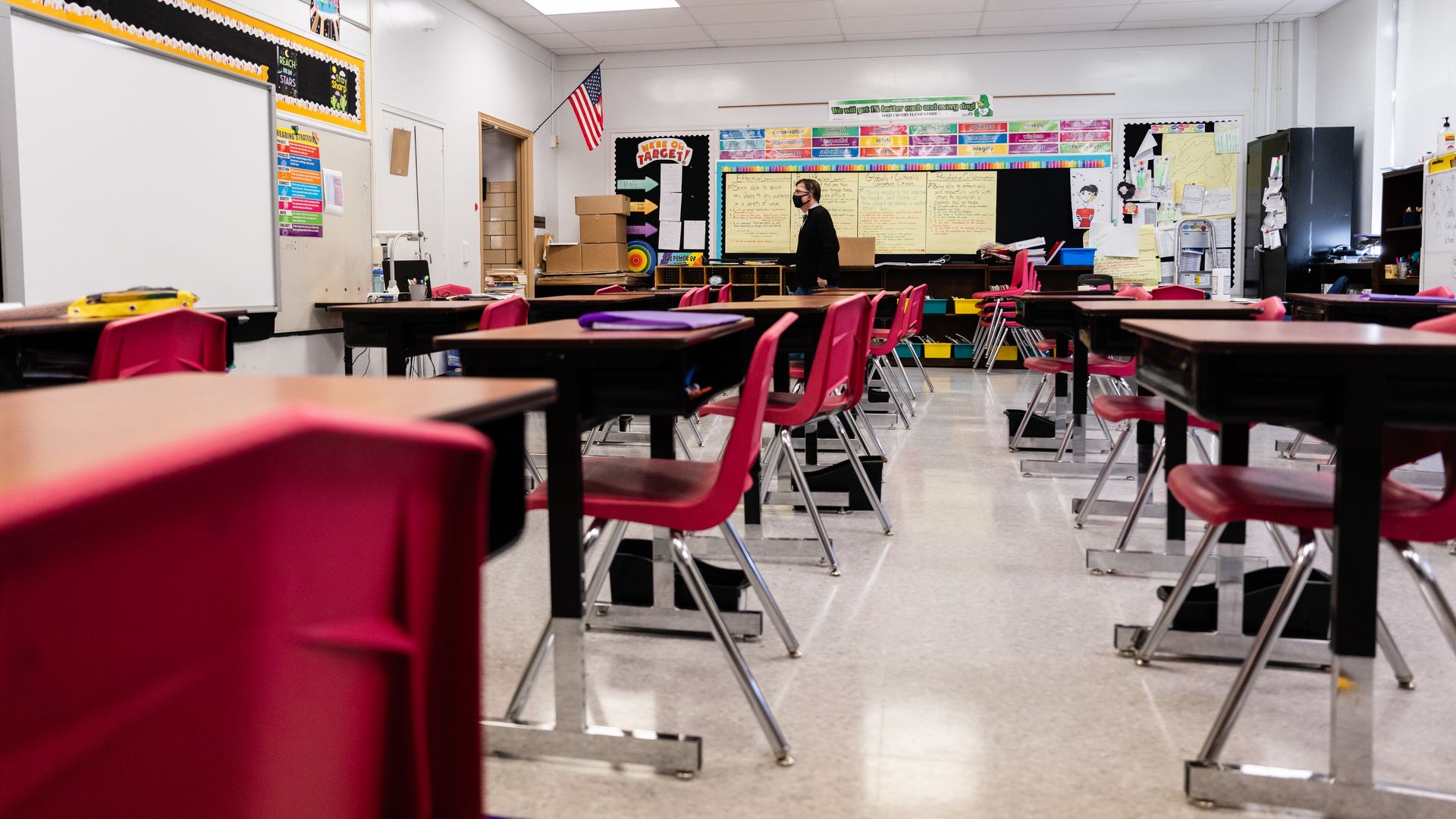 Photo of a masked person standing in the middle of a school classroom filled with desks and red chairs