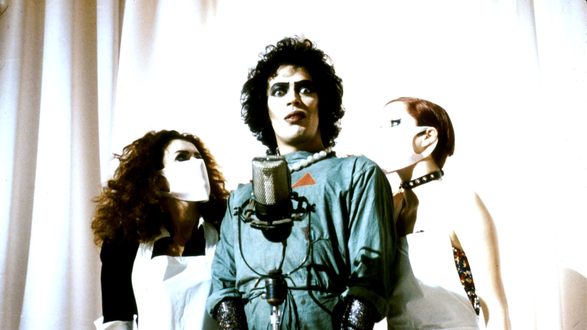 A scene from the Rocky Horror Picture show with Tim Curry's Dr. Frank-N-Furter in front of a microphone with two "nurses" by his side.