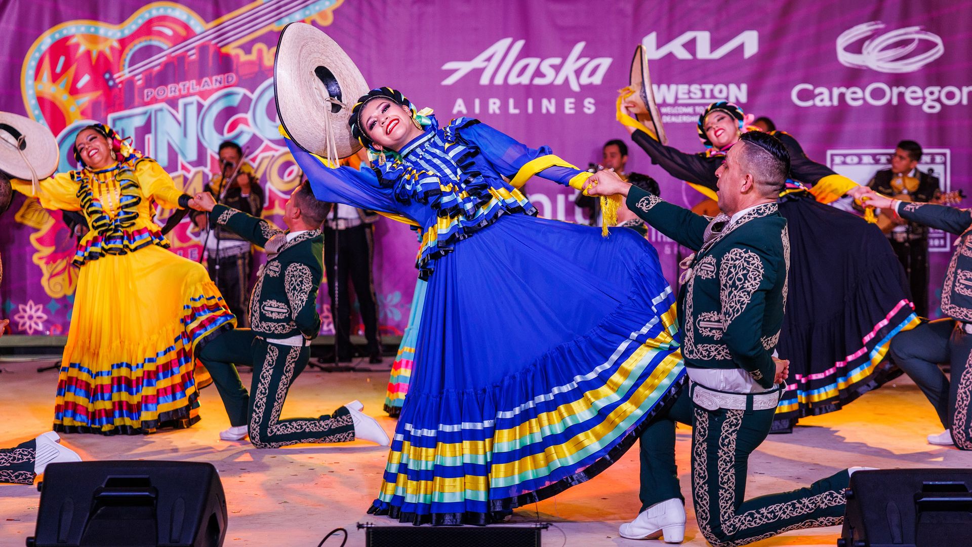 Dancers in colorful traditional Mexican attire perform a folkloric dance at Portland's Cinco de Mayo festival, holding sombreros and smiling as mariachi musicians play in the background.
