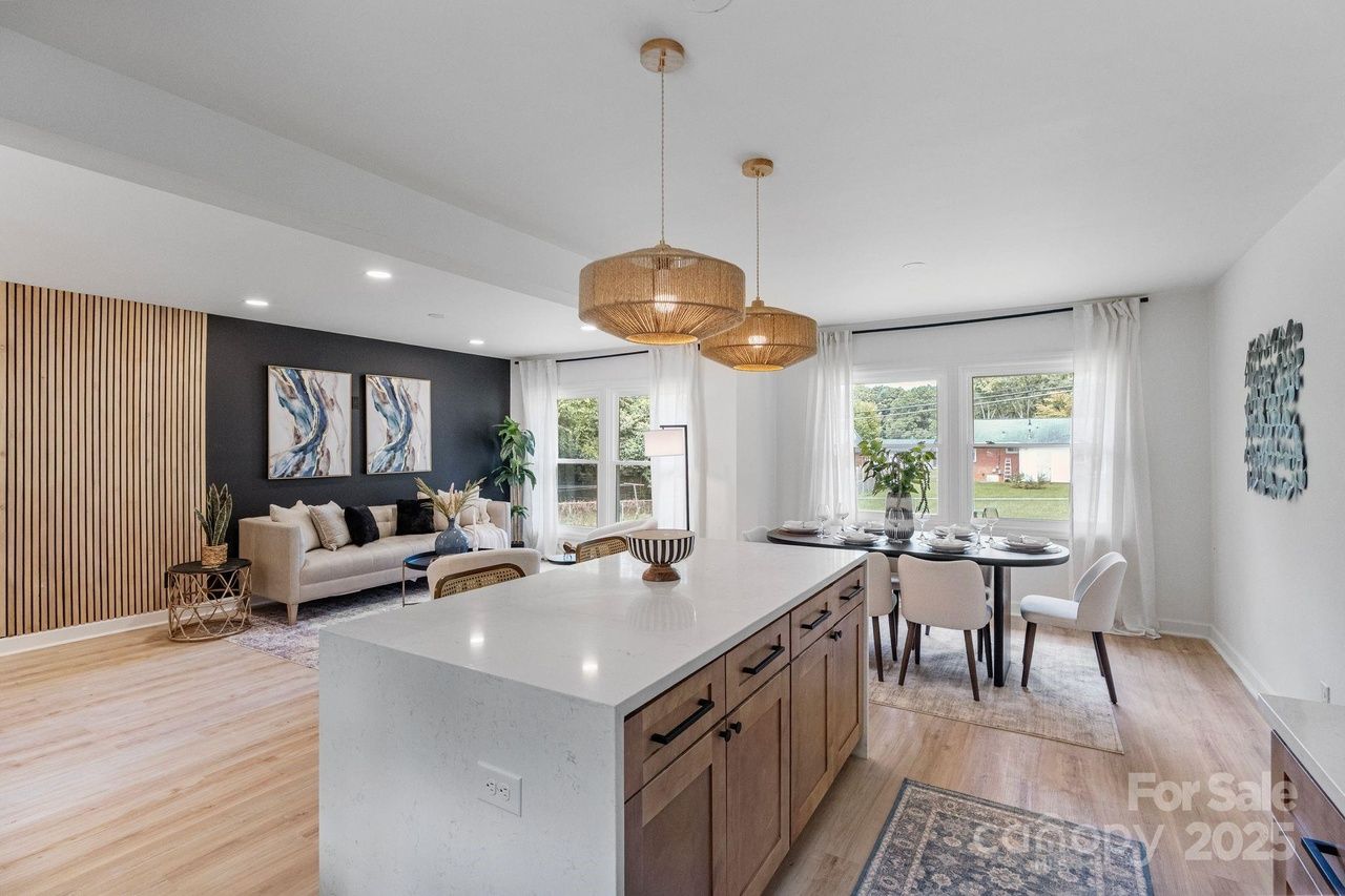 Open living and dining area with light wood floors, beige sofa, black accent wall with abstract art, white curtains, wooden island with white countertop, and round dining table with white chairs.