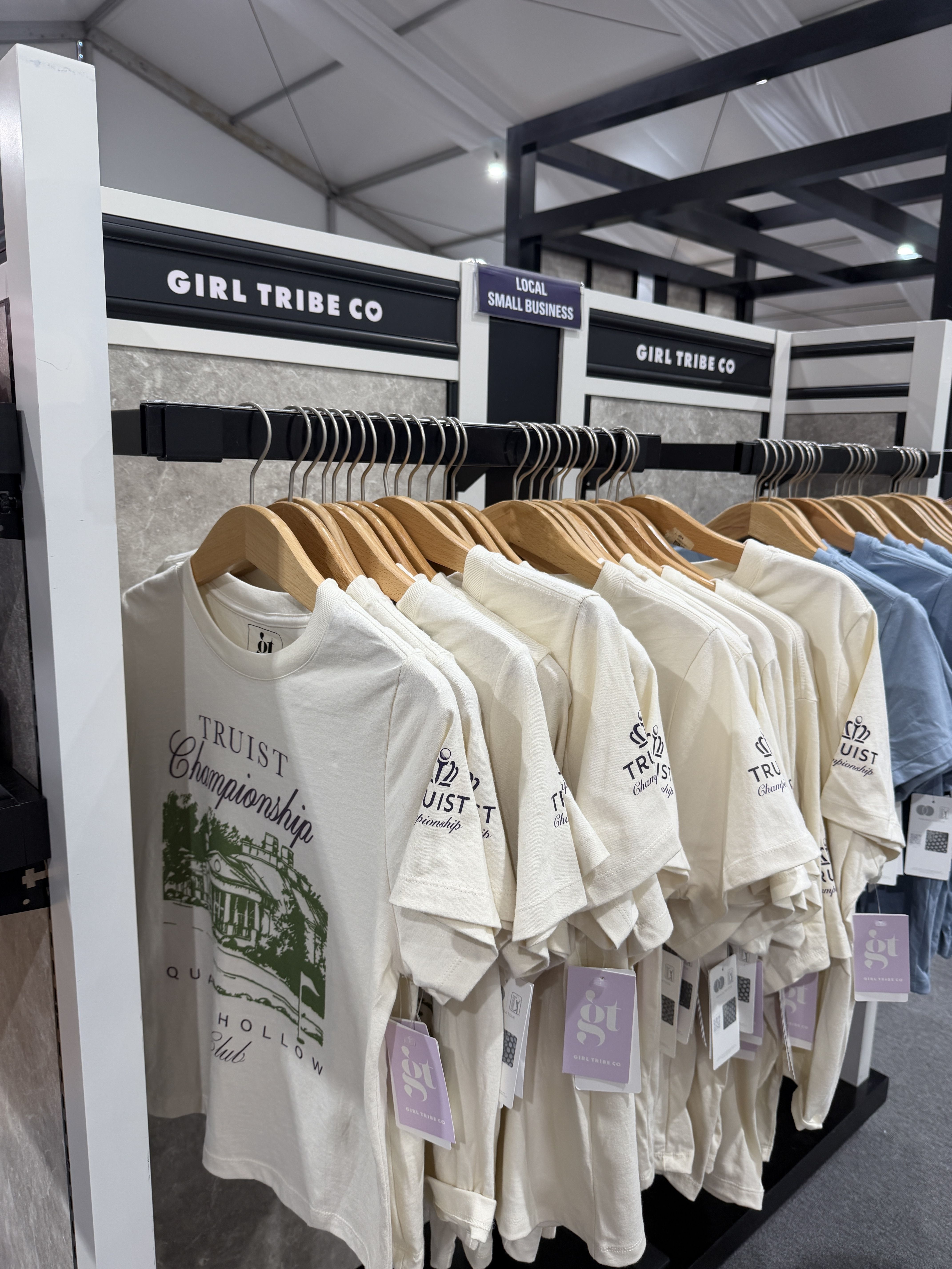 Beige T-shirts on wooden hangers along a black display rack in a store, with signs reading "GIRL TRIBE CO" and a small overhead banner that says "LOCAL SMALL BUSINESS."