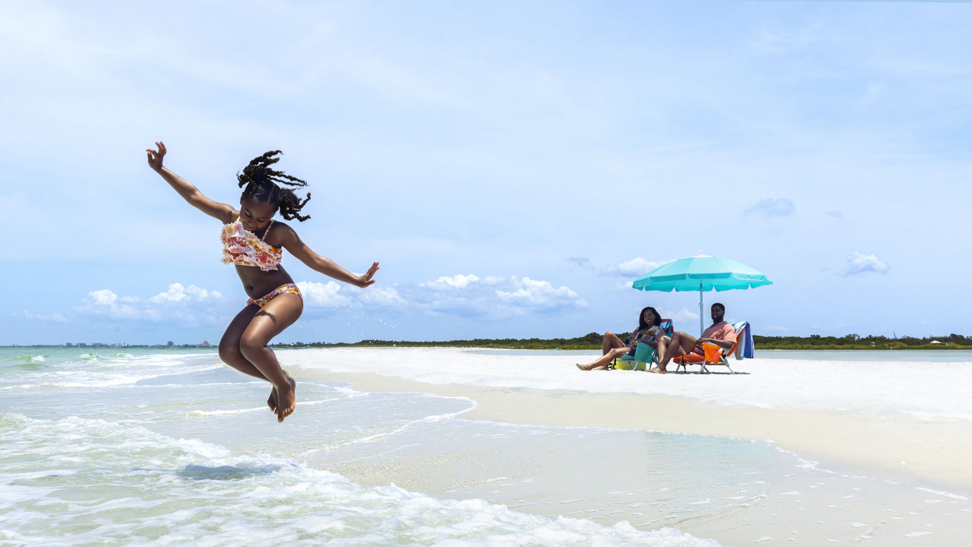 Young girl in a floral bikini leaps into the shallow surf at a sunny beach; in the background, two people lounge under a turquoise umbrella on white sand beneath a bright blue sky.