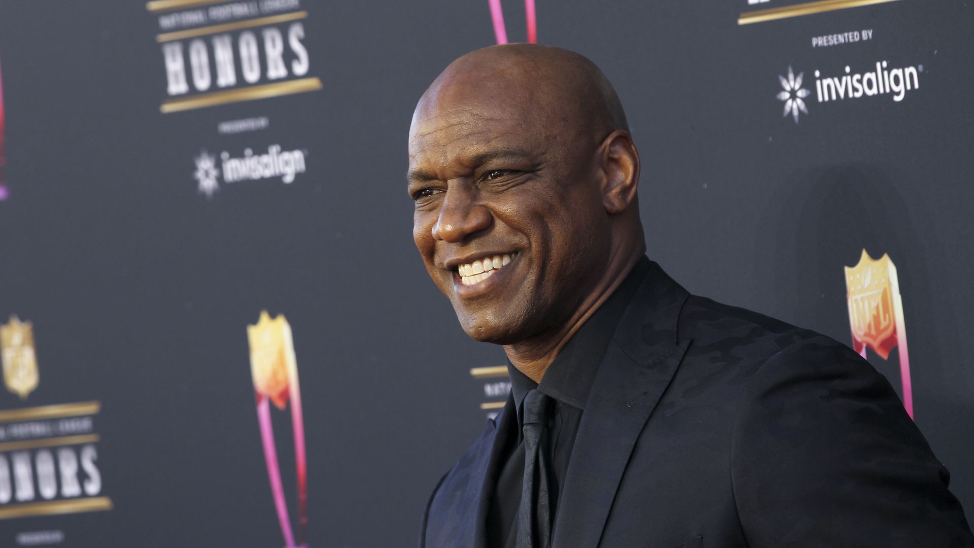 Close-up of a smiling, bald Black man in a black suit on a step-and-repeat backdrop with logos reading "HONORS" and "Invisalign".
