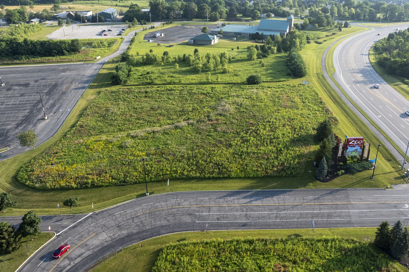 Aerial view of a grassy field with wildflowers, bordered by a curved multi-lane road on the right and the Columbus Zoo's parking lot on the left; a zoo sign sits near the foreground.