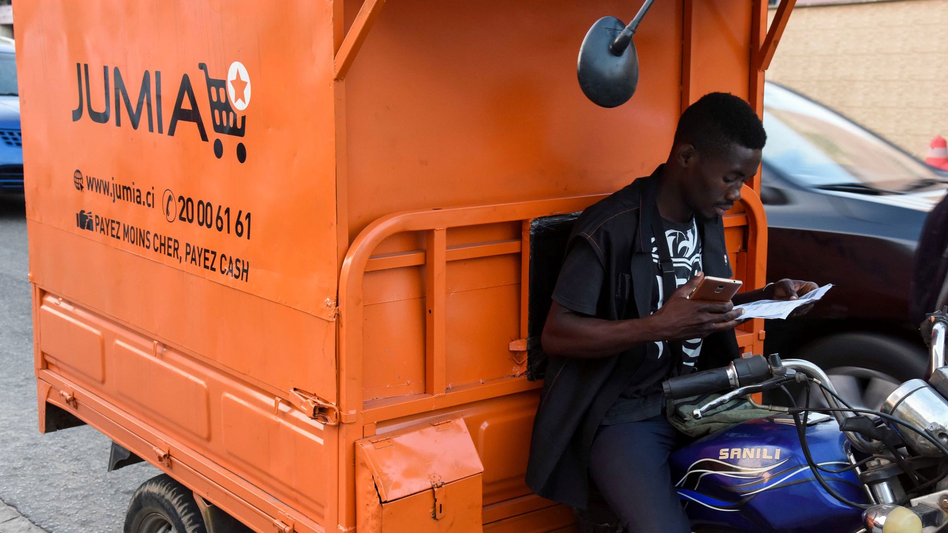 A man studies a piece of paper while leaning against an orange Jumia delivery vehicle