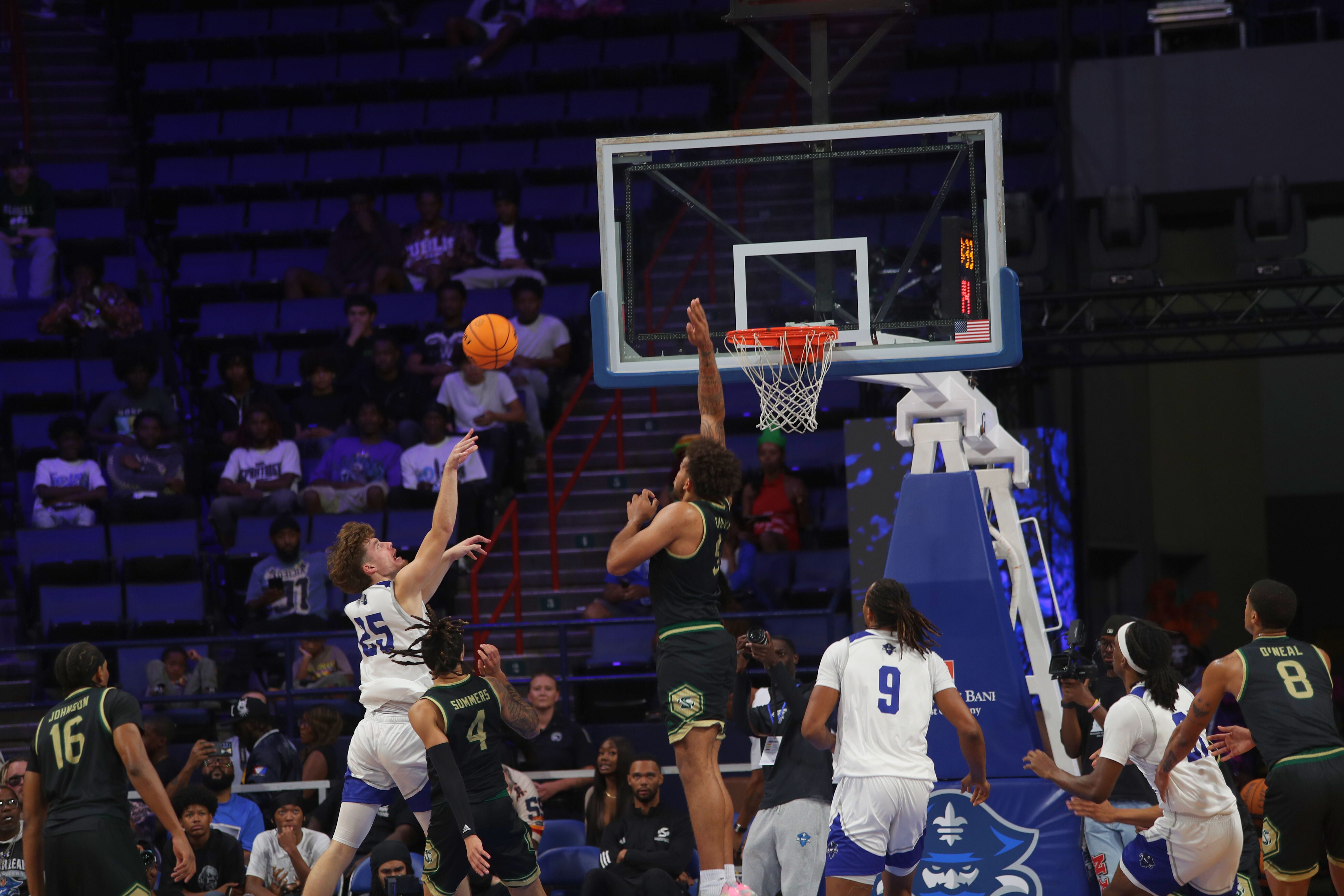 Basketball game showing player 25 in white shooting with defense player 4 in black jumping to block under the hoop, with other players and spectators in a purple-seated arena.