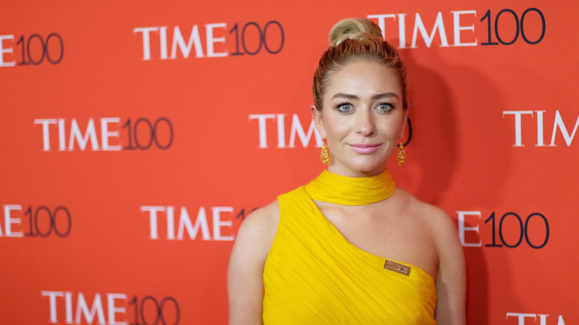 A blonde woman in a yellow dress stands in front of a photo wall with the TIME100 logo in a pattern